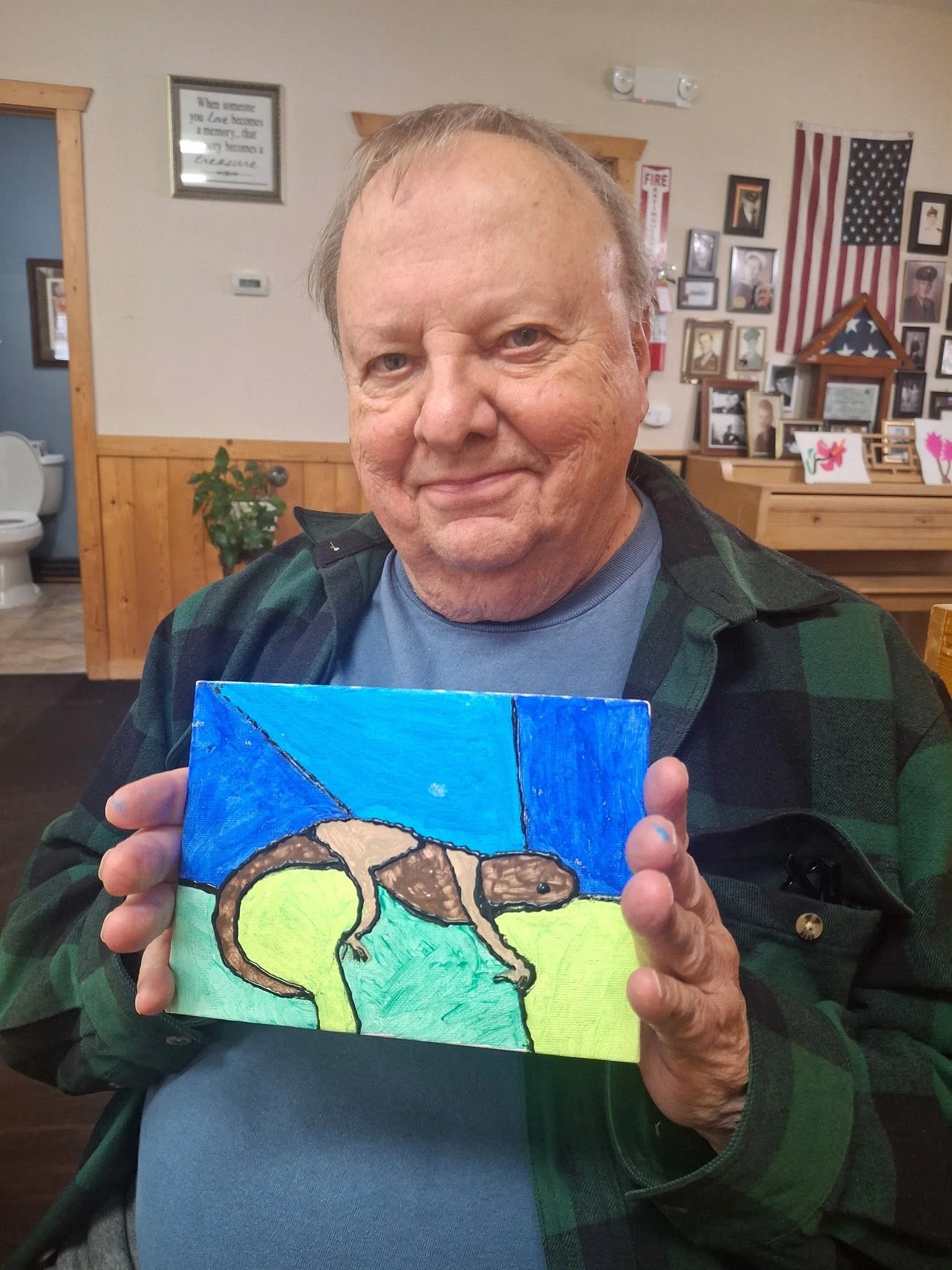 An older man with a bald head and gray hair on the sides, smiling, holding a colorful painting of a squirrel on a branch, with background colors of blue, green, and yellow.