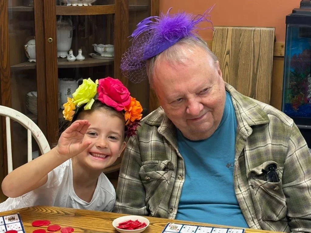 A young girl and an older man sitting at a table. The girl is smiling and waving, wearing a colorful floral headband. The man is wearing a purple feathered hat and a plaid shirt. There are red game pieces and cards on the table.