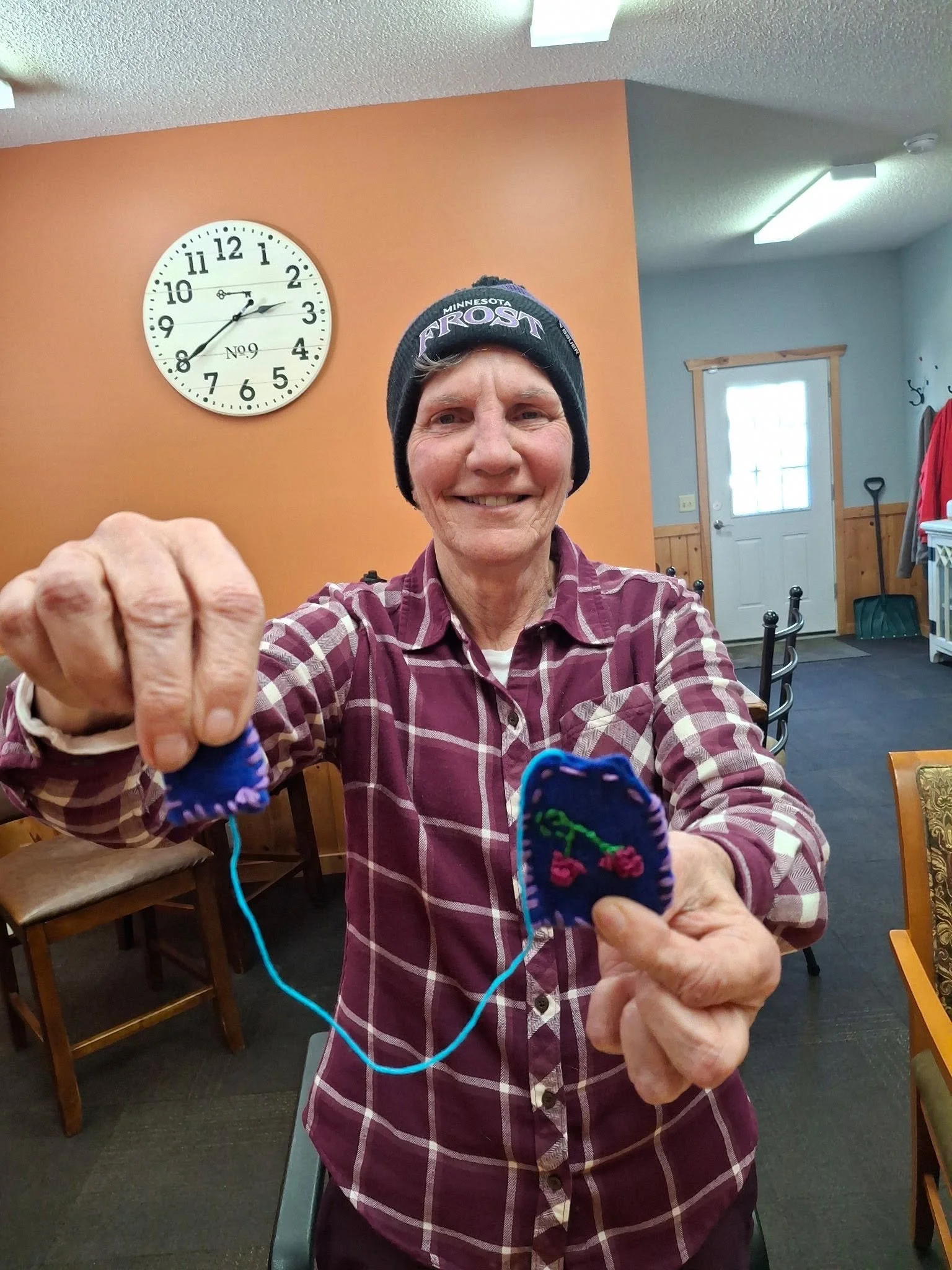 An elderly woman smiling and holding knitting needles with blue yarn and colorful socks. She is wearing a checkered shirt and a headband that says Minnesota Frost.
