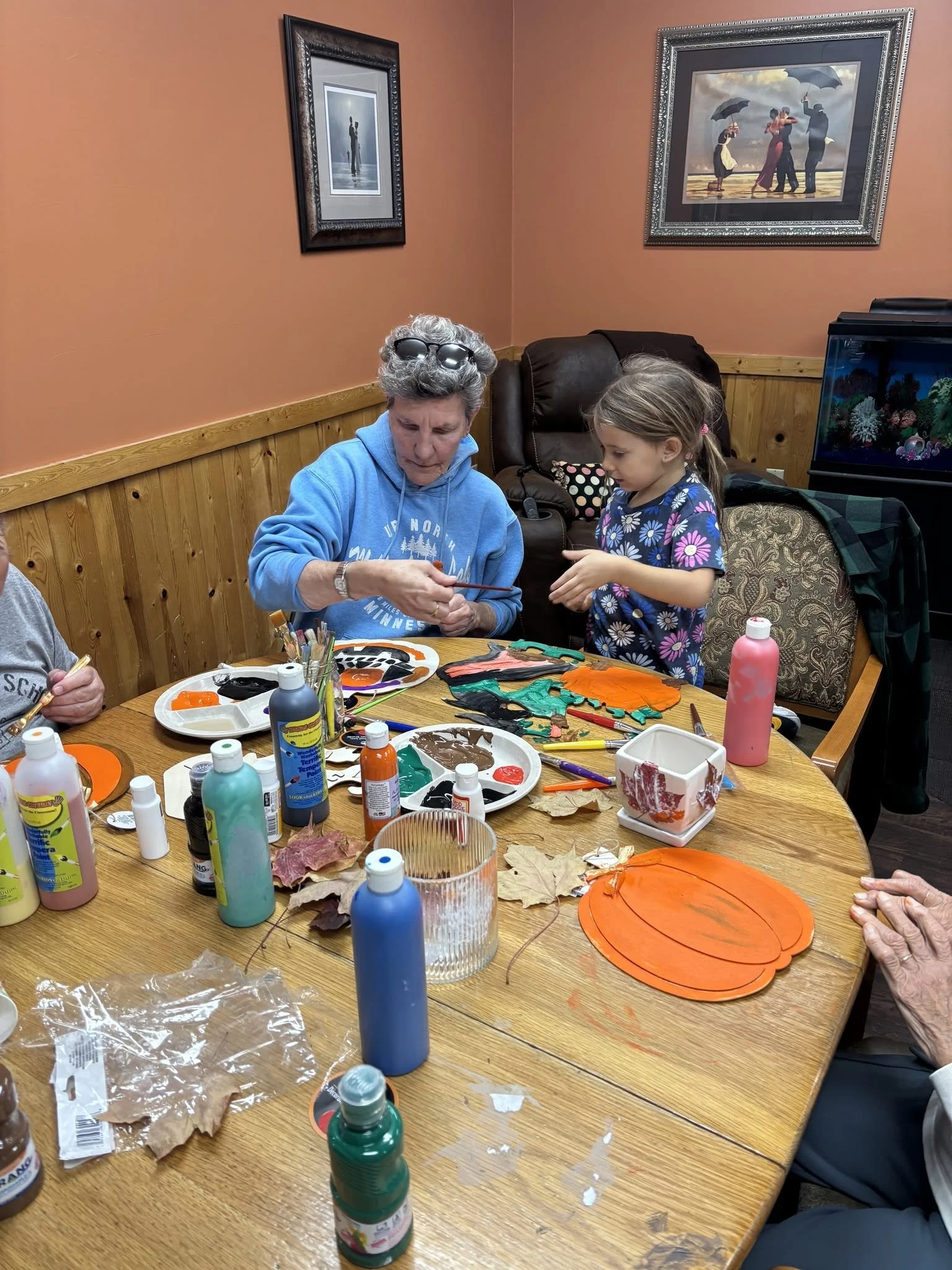 A woman with gray hair and sunglasses on her head, and a young girl with a ponytail, are working on a craft project at a round wooden table in a room with peach walls. The table is filled with art supplies, painted paper cutouts, and bottles of paint