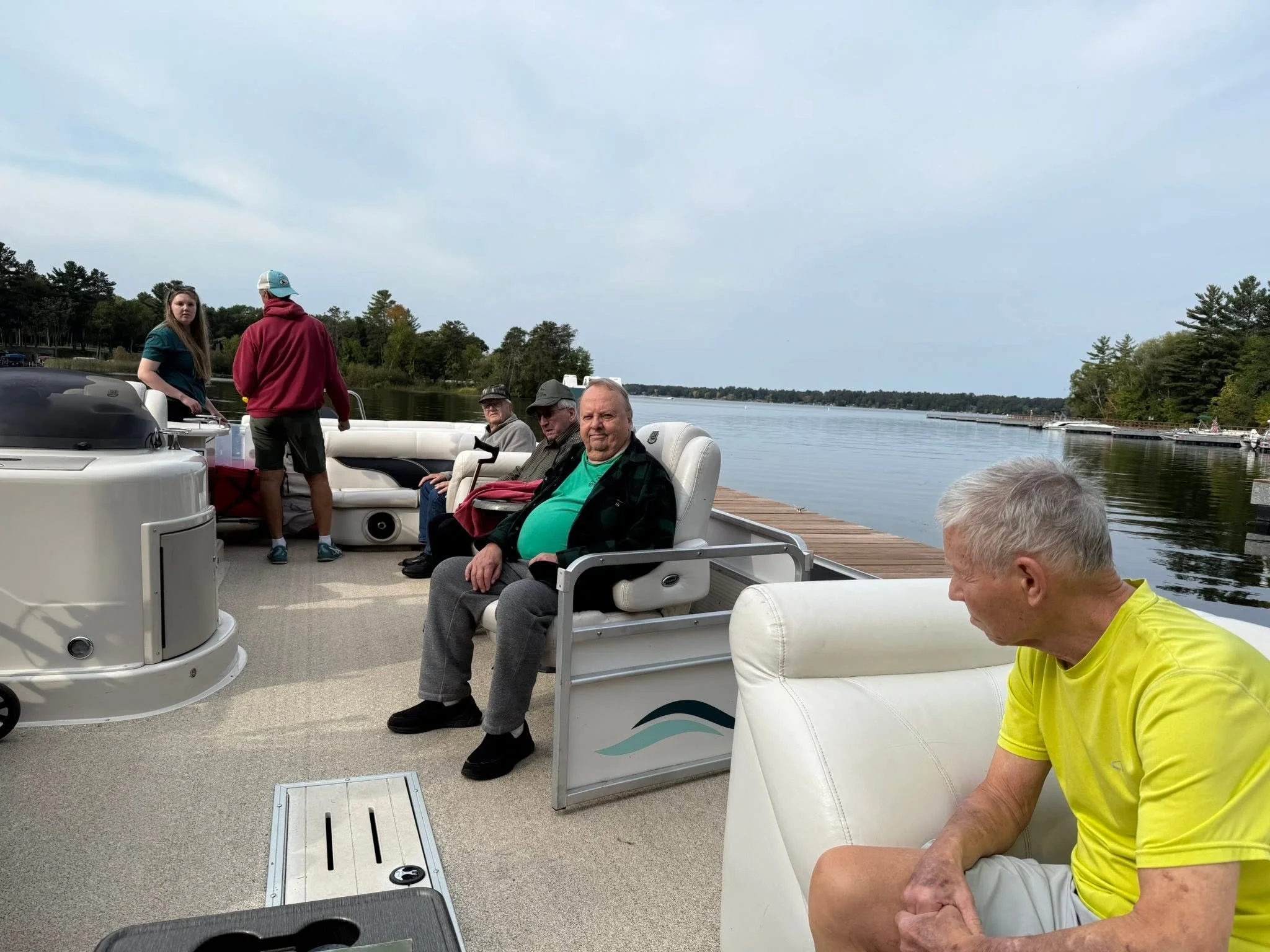 People sitting and standing on a boat docked in a lake, with trees and water in the background, some individuals are wearing casual clothes and hats, and appear to be enjoying a boat trip.