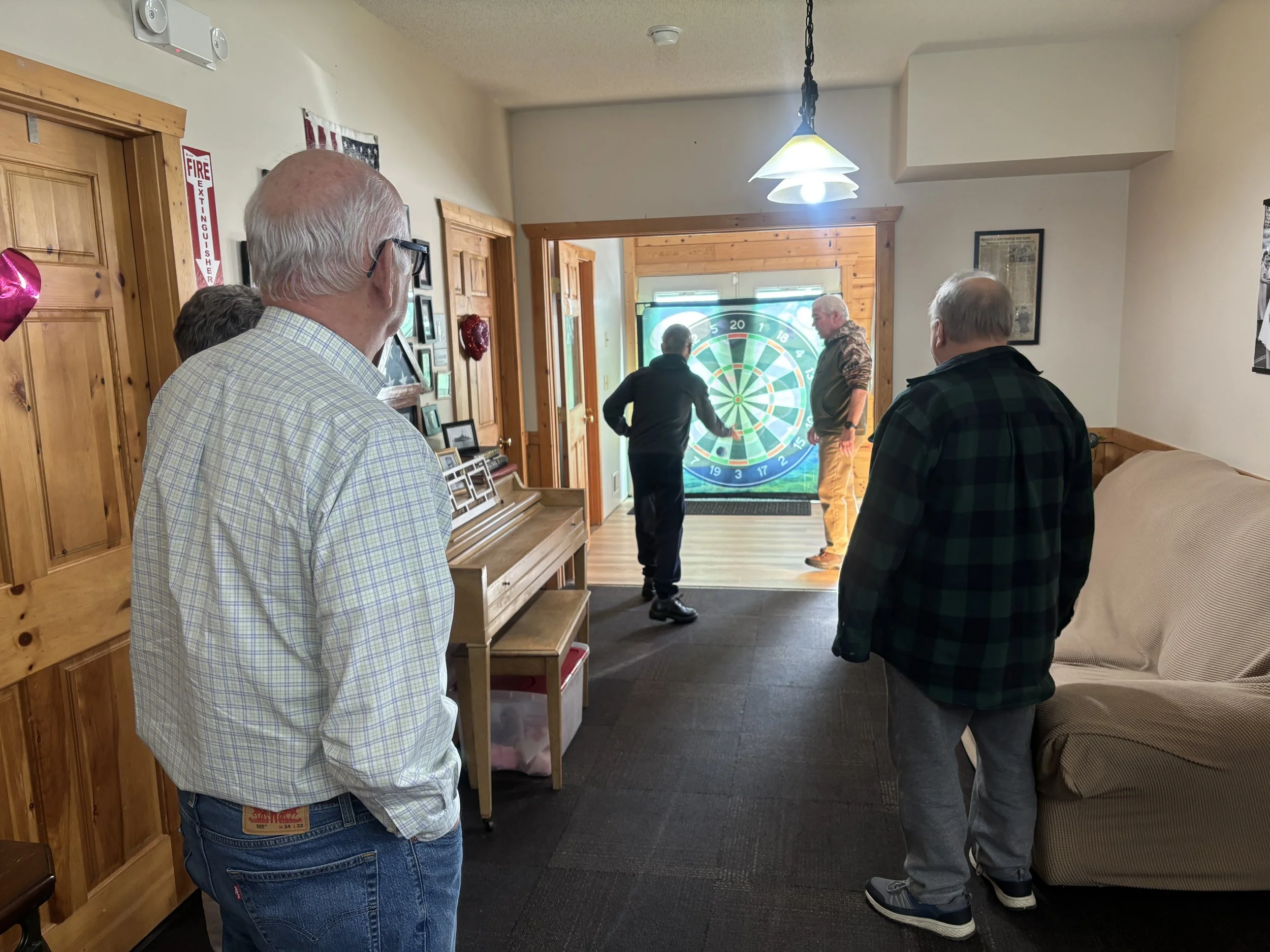 A group of people gather in a room with a dartboard displayed on a screen at the front. The room has wood-paneled walls and framed pictures, with some individuals watching the game while others participate.