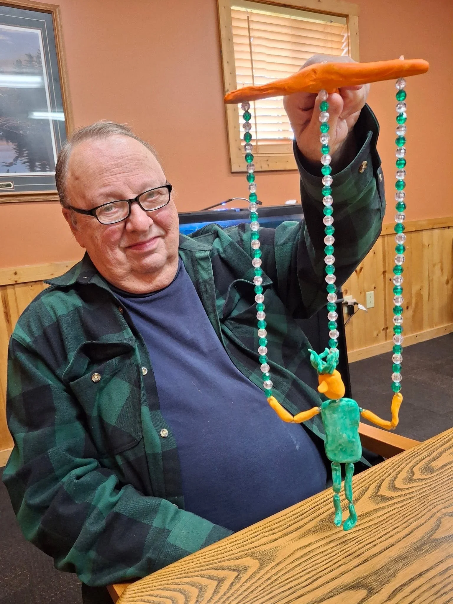 An elderly man with glasses is sitting at a wooden table, smiling at the camera. He is wearing a dark blue shirt and a green plaid jacket. He is holding up a colorful beaded sculpture resembling an insect or spider, made of green, transparent, and or