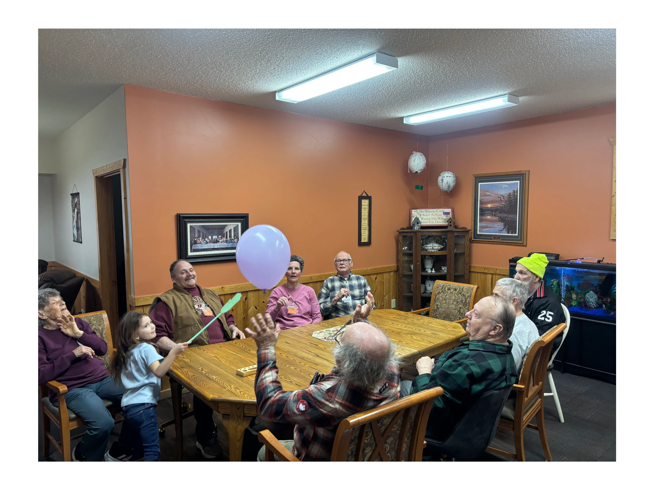 Group of people seated around a wooden table in a room with orange walls, decorated with framed pictures and hanging paper lanterns. Some people are smiling, and there is a purple balloon in the foreground.