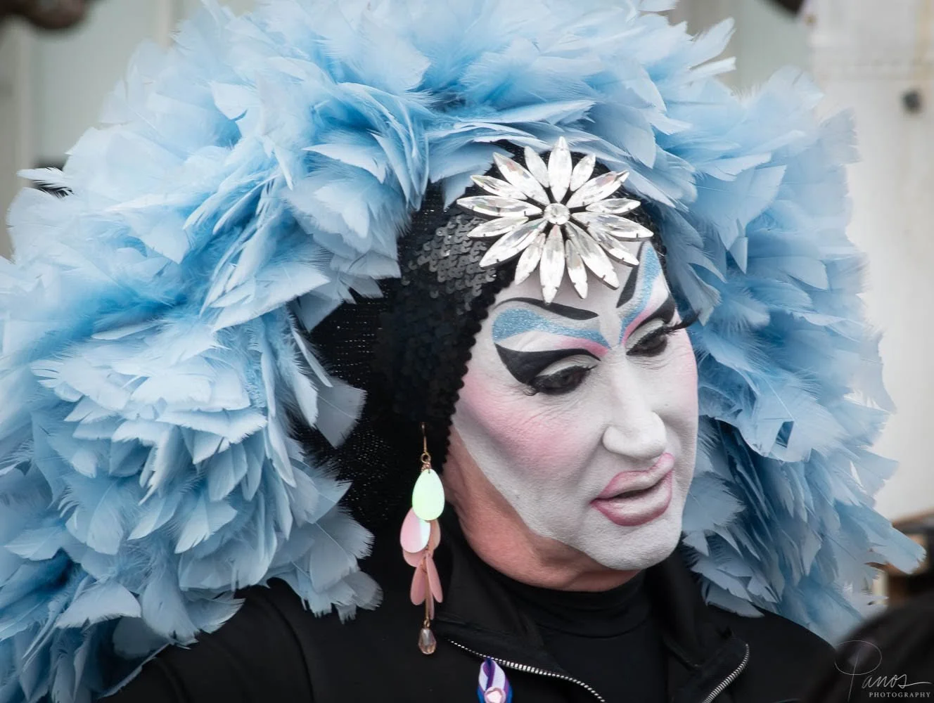 Sister of Perpetual Indulgence framed by pale blue feathers and wearing theatrical white face makeup.
