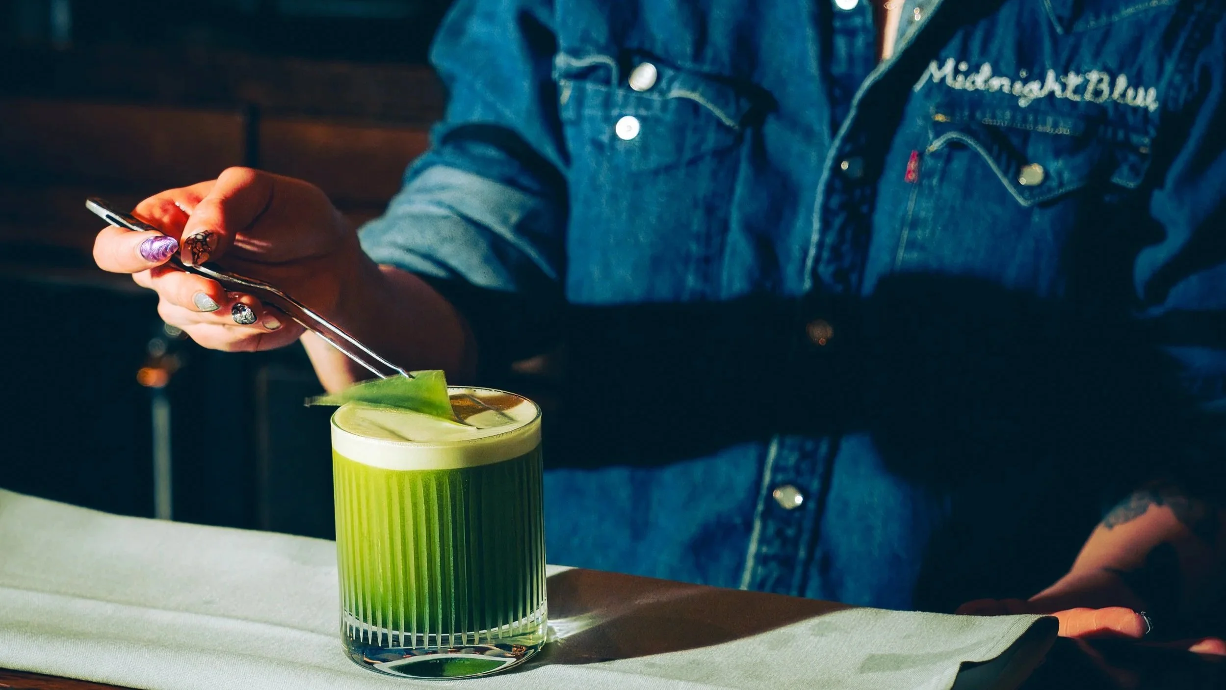 A person wearing a denim jacket with embroidery on the left chest is stirring a layered green and white cocktail with a green leaf garnish.