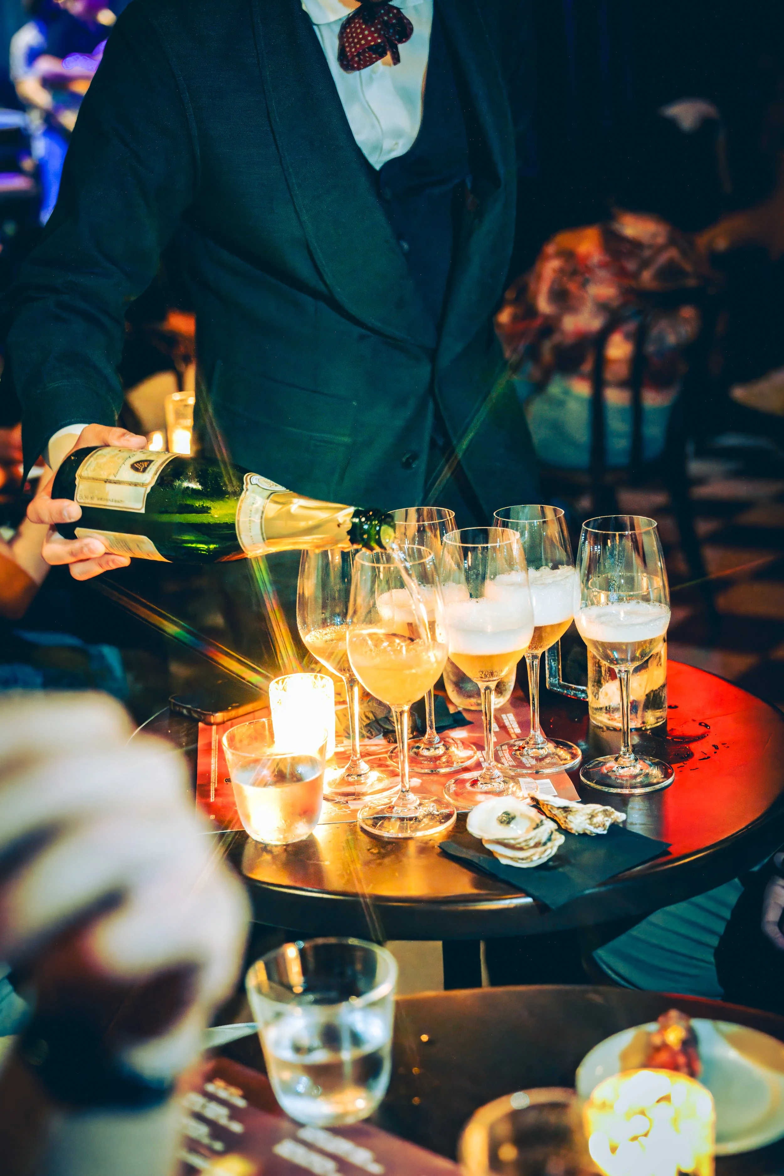 Server pouring champagne into glasses at a bar or restaurant setting, with oysters on a napkin on the table, illuminated by candlelight.