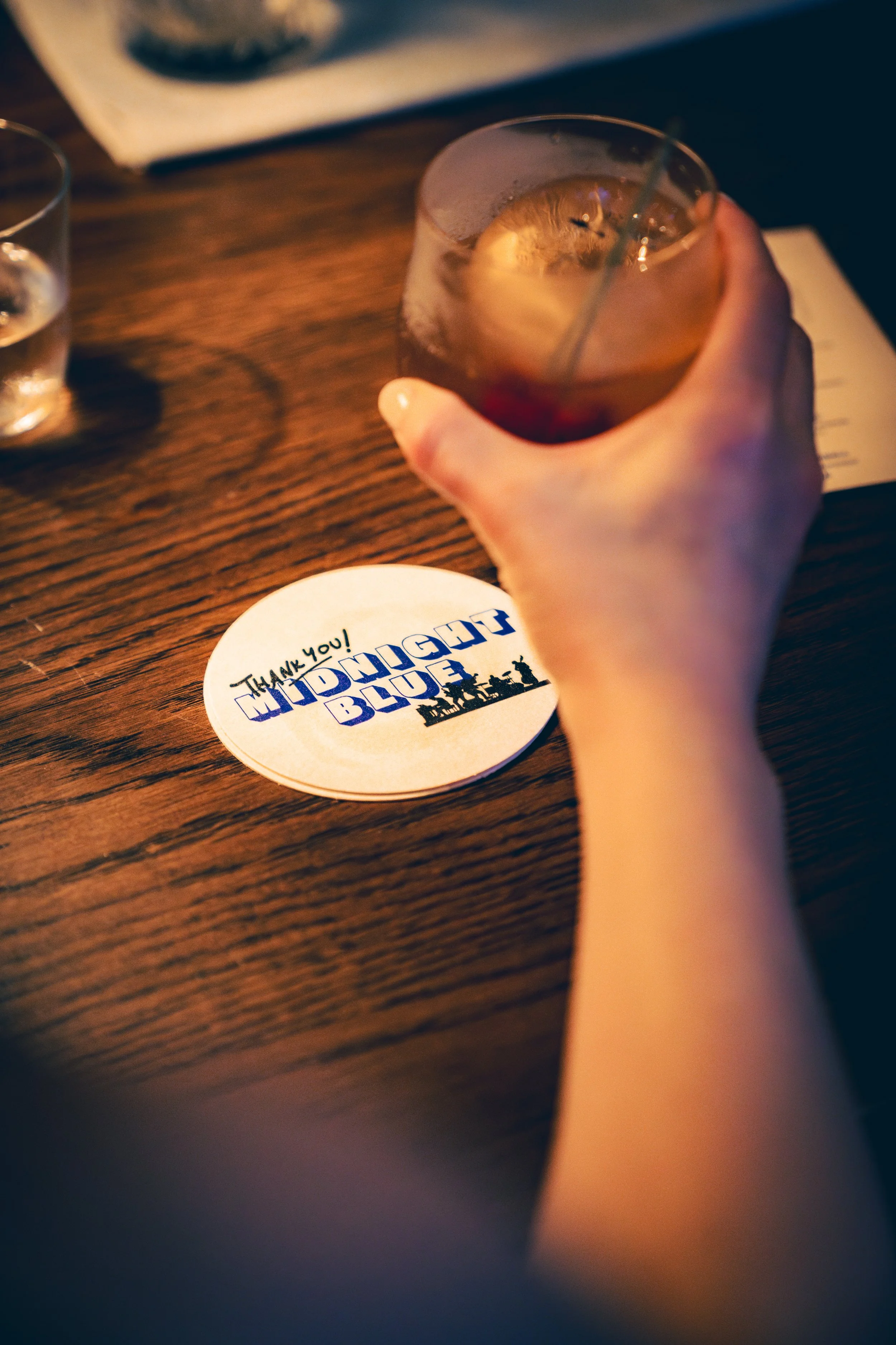 Person holding a drink in a glass with a straw on a wooden table, next to a coaster with the text 'Thank you! Midnight Blue' and a silhouette of city buildings.