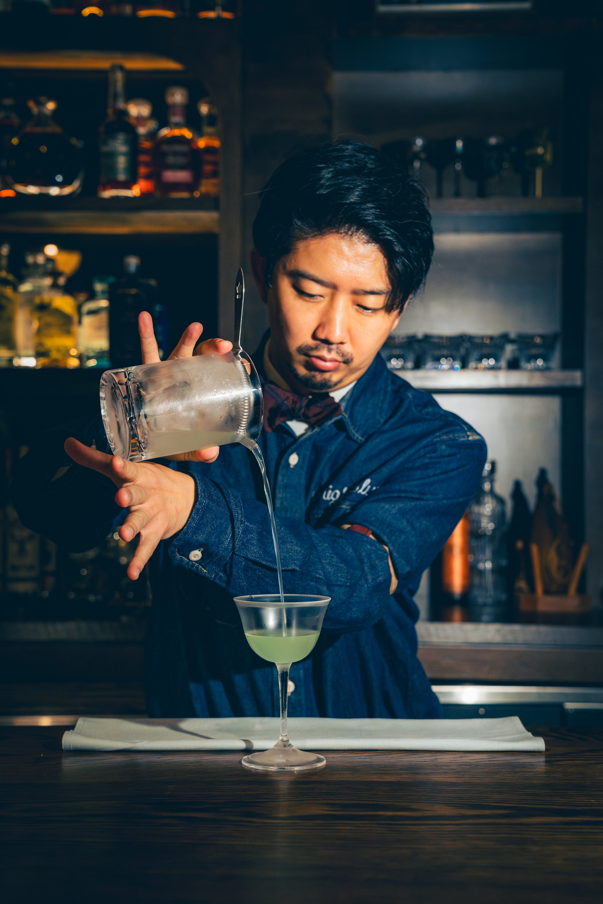 Bartender pouring a clear liquid into a green cocktail in a martini glass behind a bar