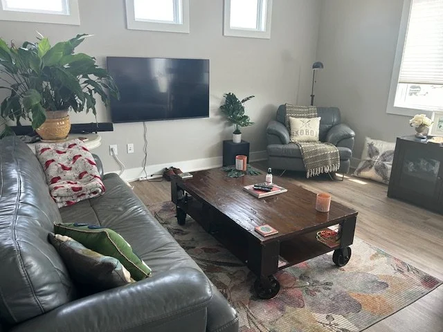 Living room with a gray leather sofa, a wooden coffee table, a gray armchair, a wall-mounted TV, and various plants and decor.
