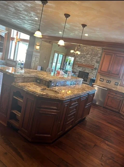 A spacious kitchen with a large granite island, wooden cabinets, a stone fireplace, and dining area with a chandelier, illuminated by natural light from large windows.