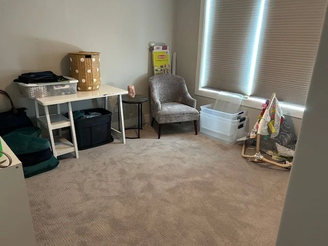 A small, organized room with a beige carpet, a white desk with storage containers, a polka-dot laundry hamper, a gray upholstered chair, a small black side table, clear plastic storage bins, and a partially opened window with closed blinds.