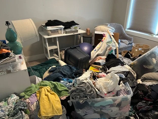 A cluttered room with clothes, bags, and miscellaneous items scattered across furniture and the floor, including a plastic storage bin filled with laundry and an exercise ball.