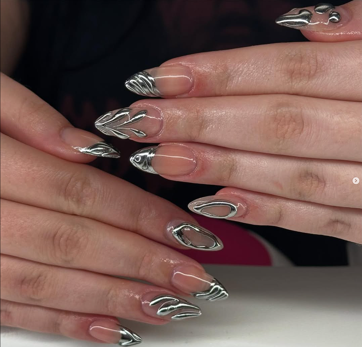 Close-up of hands with long, almond-shaped nails featuring metallic silver abstract designs on clear nail tips.