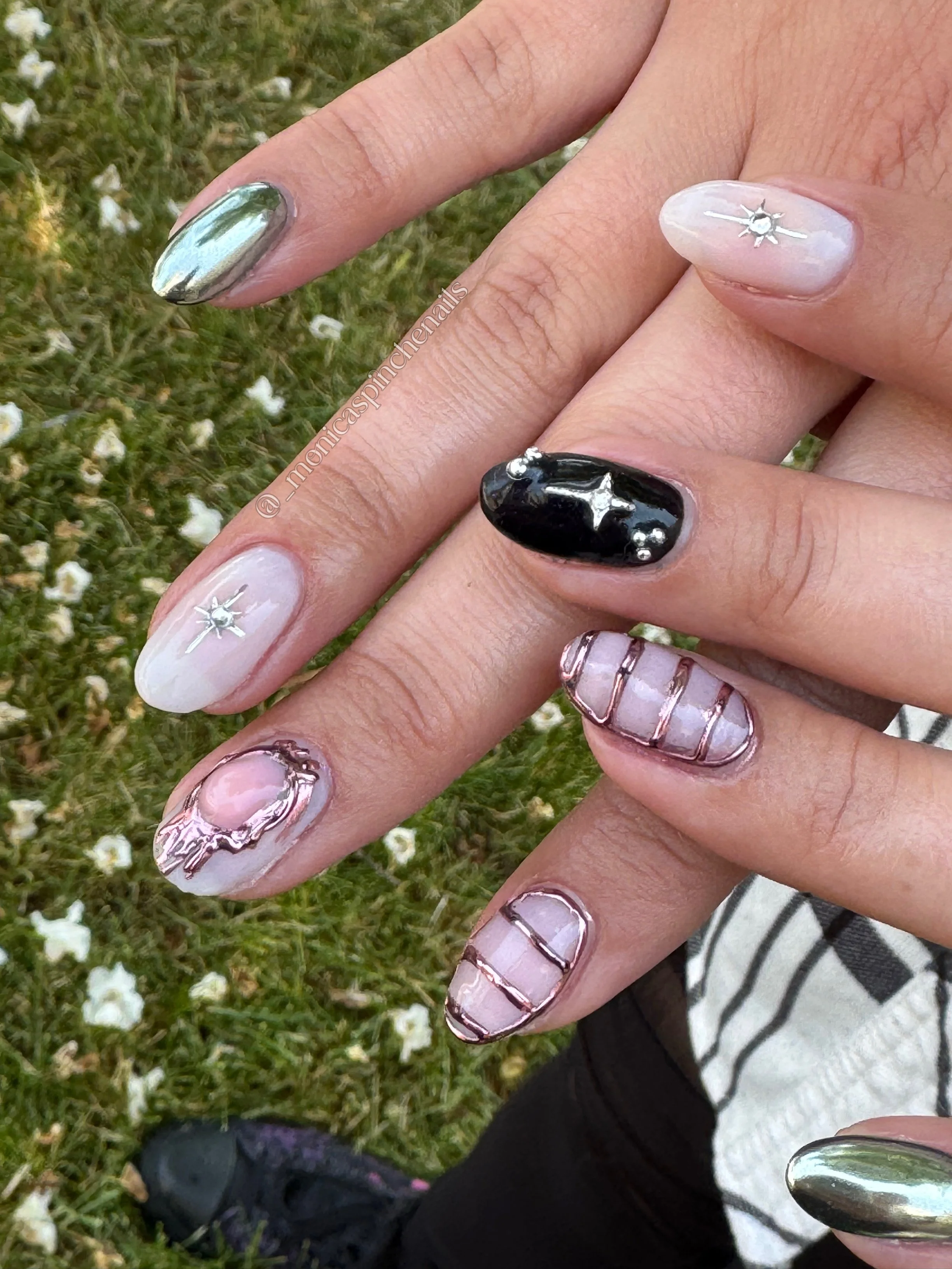 Close-up of a hand with various nail art designs on each fingernail, including metallic, black with stars, and striped patterns, held above grass with small white flowers.