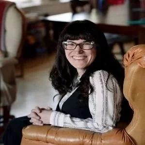 A woman with black hair and glasses smiling while seated in a cozy casual setting.