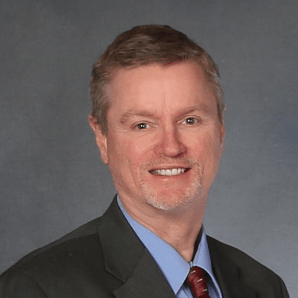 A professional headshot of Roy Neese, a man with light brown hair and a goatee, wearing a dark suit, blue shirt, and red tie, smiling against a neutral background.