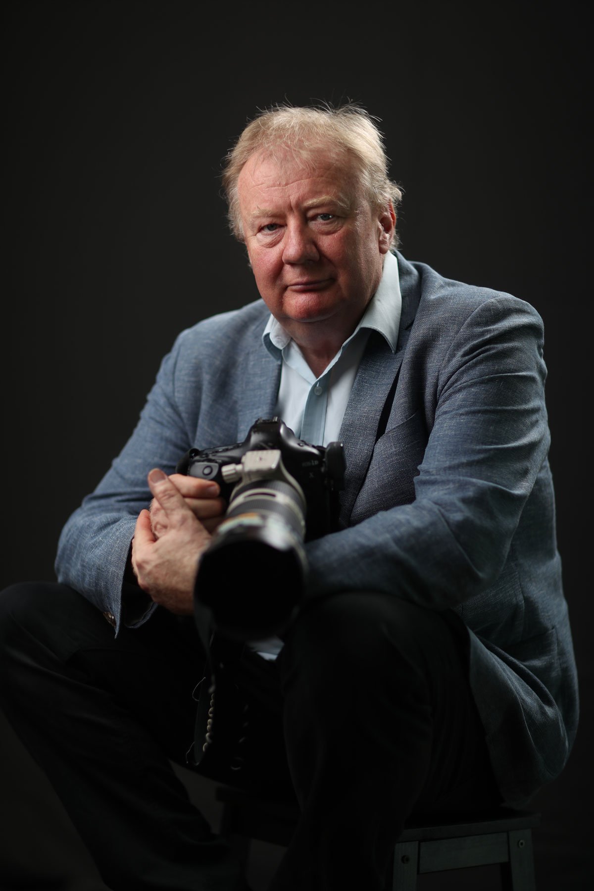 An older man with light-colored hair sits on a stool against a dark background, holding a professional camera with a large lens.