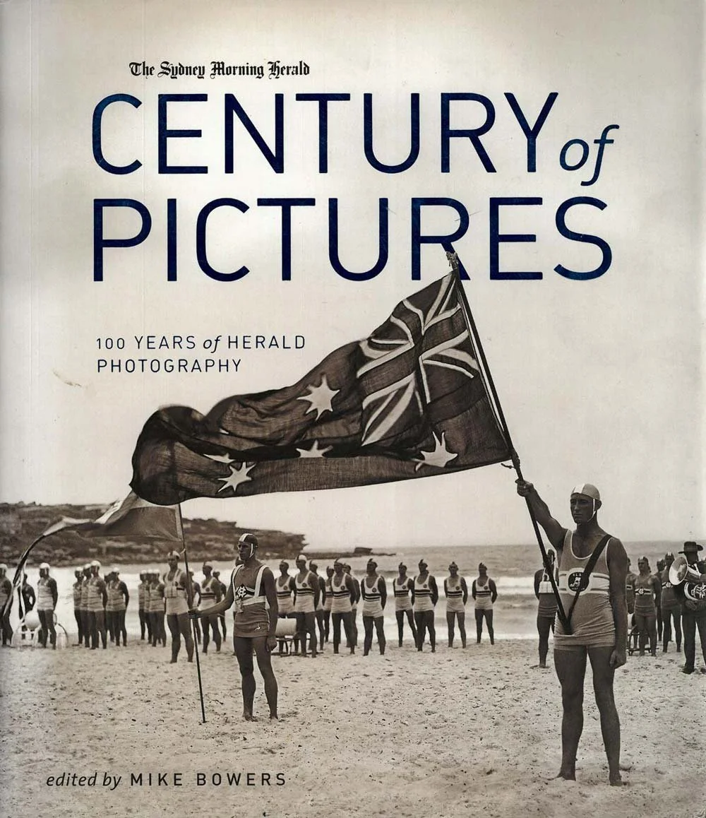 Book cover titled 'Century of Pictures: 100 Years of Herald Photography,' featuring a black-and-white photo of men at the beach, some holding Australian flags, with the ocean and a cliff in the background.