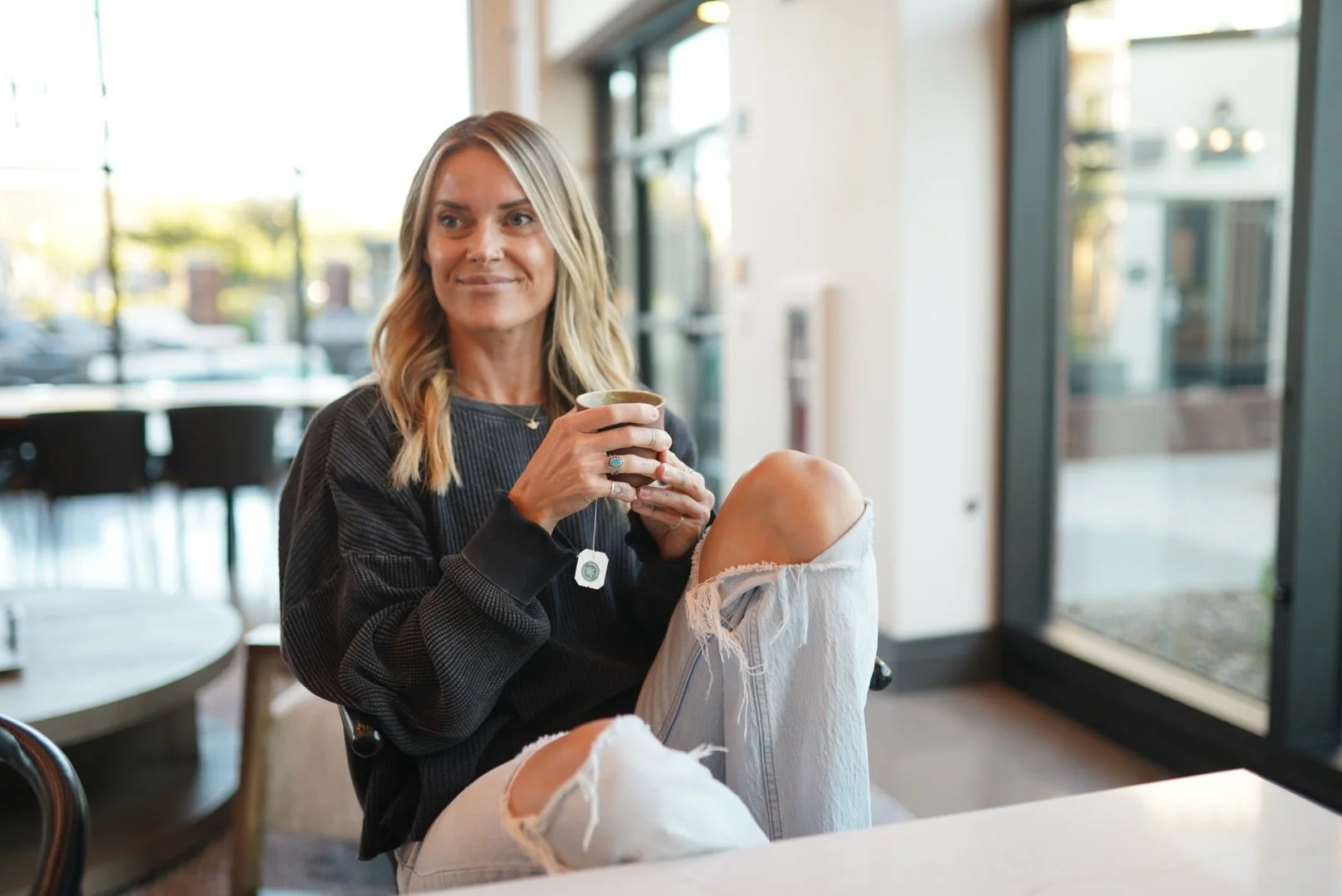 A woman with blonde hair sitting inside a cafe, holding a coffee mug, wearing a black sweater and ripped light-colored jeans, with sunlight coming through large windows behind her.