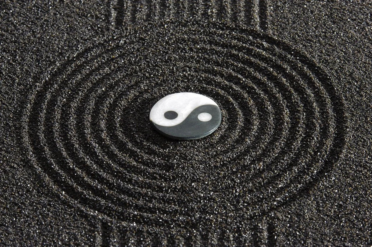 A yin-yang symbol resting on black textured sand, with concentric circular patterns surrounding it.
