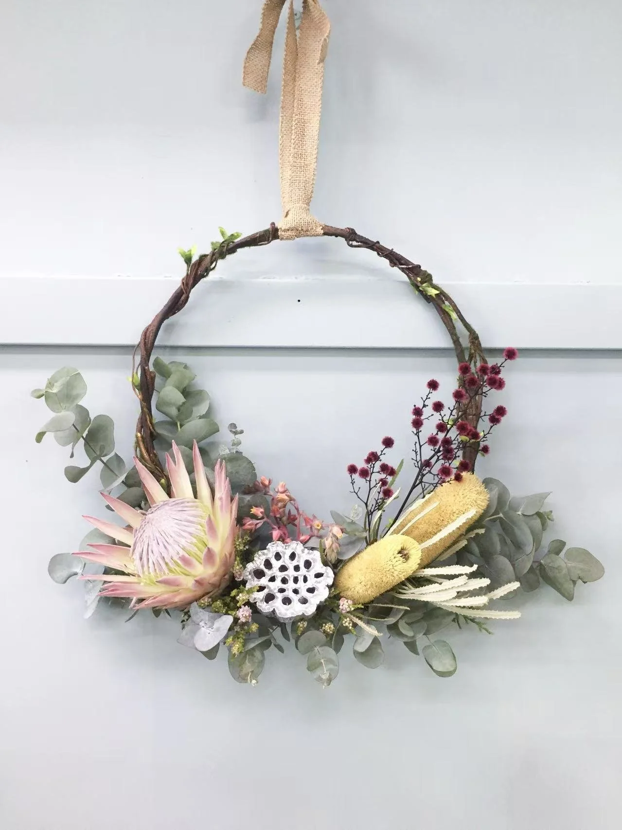 Decorative floral wreath hanging on a white wall with various flowers and greenery, including a large protea, eucalyptus leaves, and small red and yellow flowers.