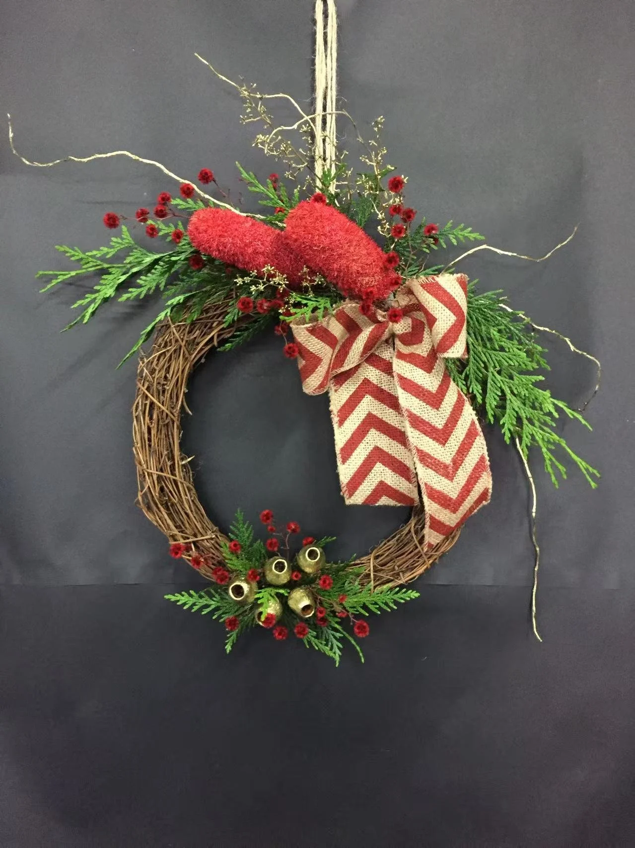 Christmas wreath with greenery, red berries, a bow, and bells hanging on a black wall.