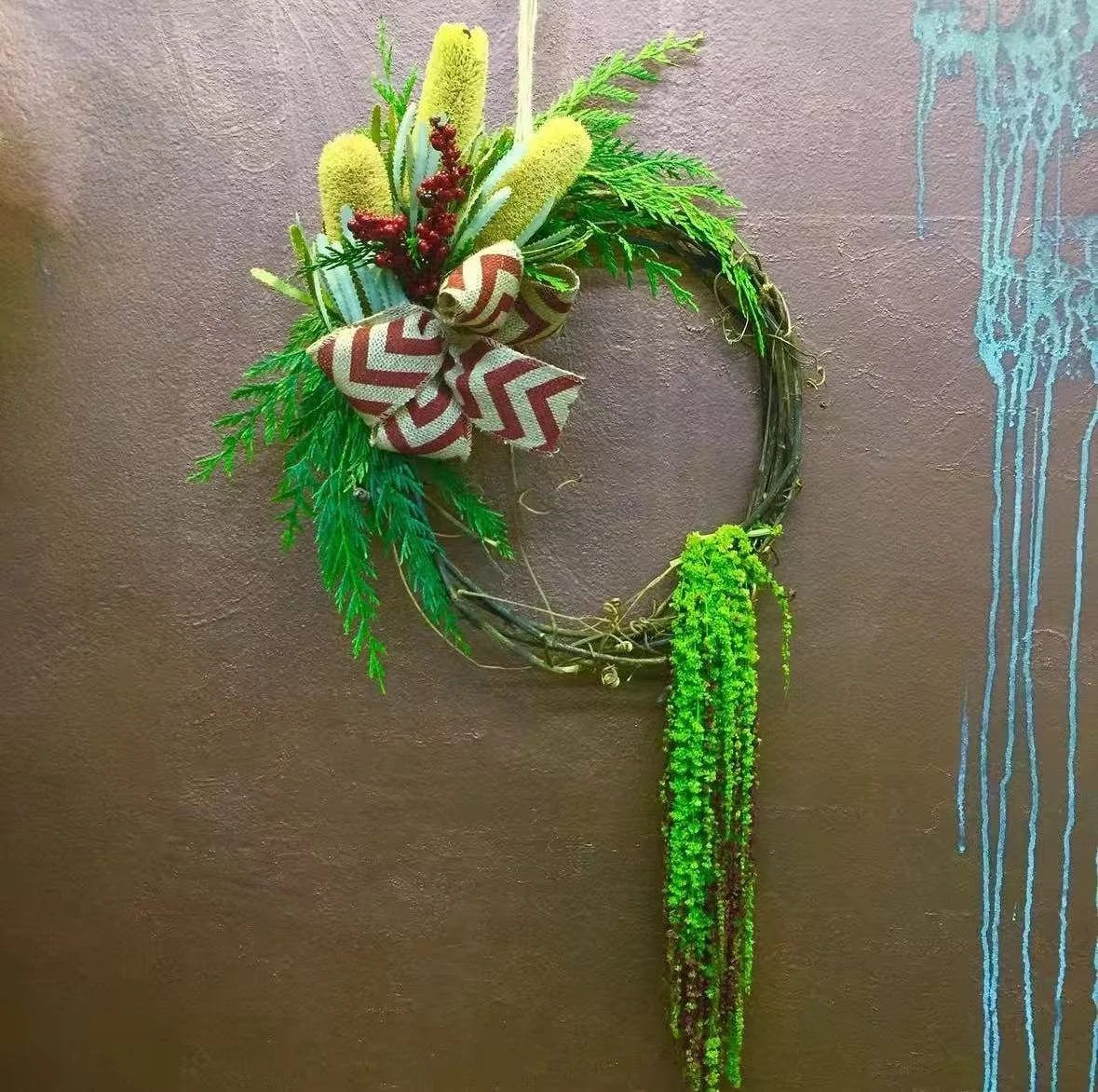 Decorative holiday wreath with green foliage, a red and white ribbon bow, yellow and red accents, mounted on a brown wall.