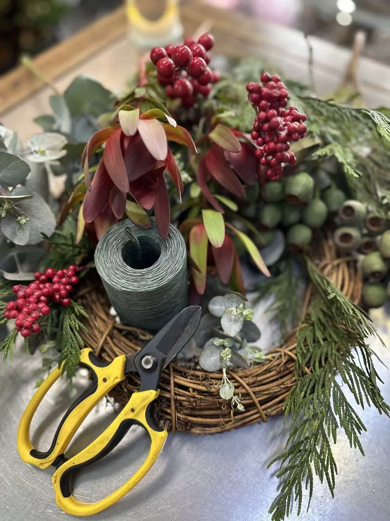 A holiday floral arrangement in a wicker basket contains red berries, green foliage, and a roll of floral tape with yellow pruning shears nearby.