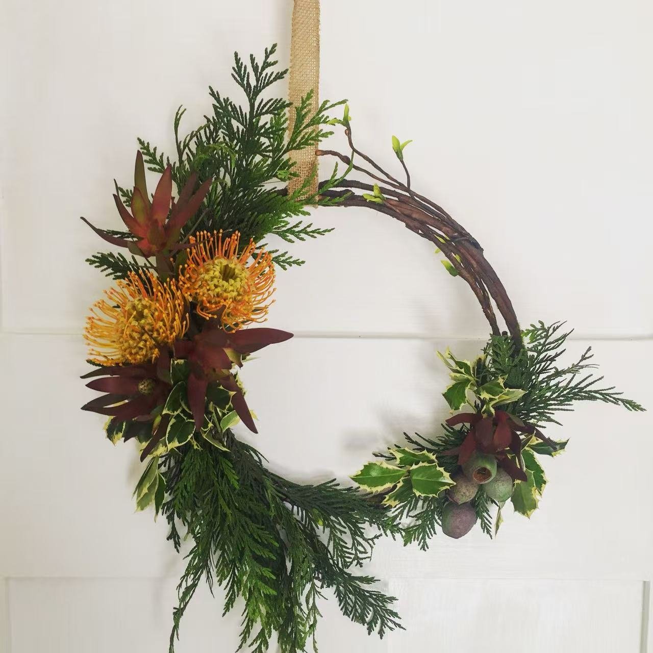A Christmas wreath made with green foliage, red flowers, yellow-orange flowers, and a branch with berries, hanging on a white wall.