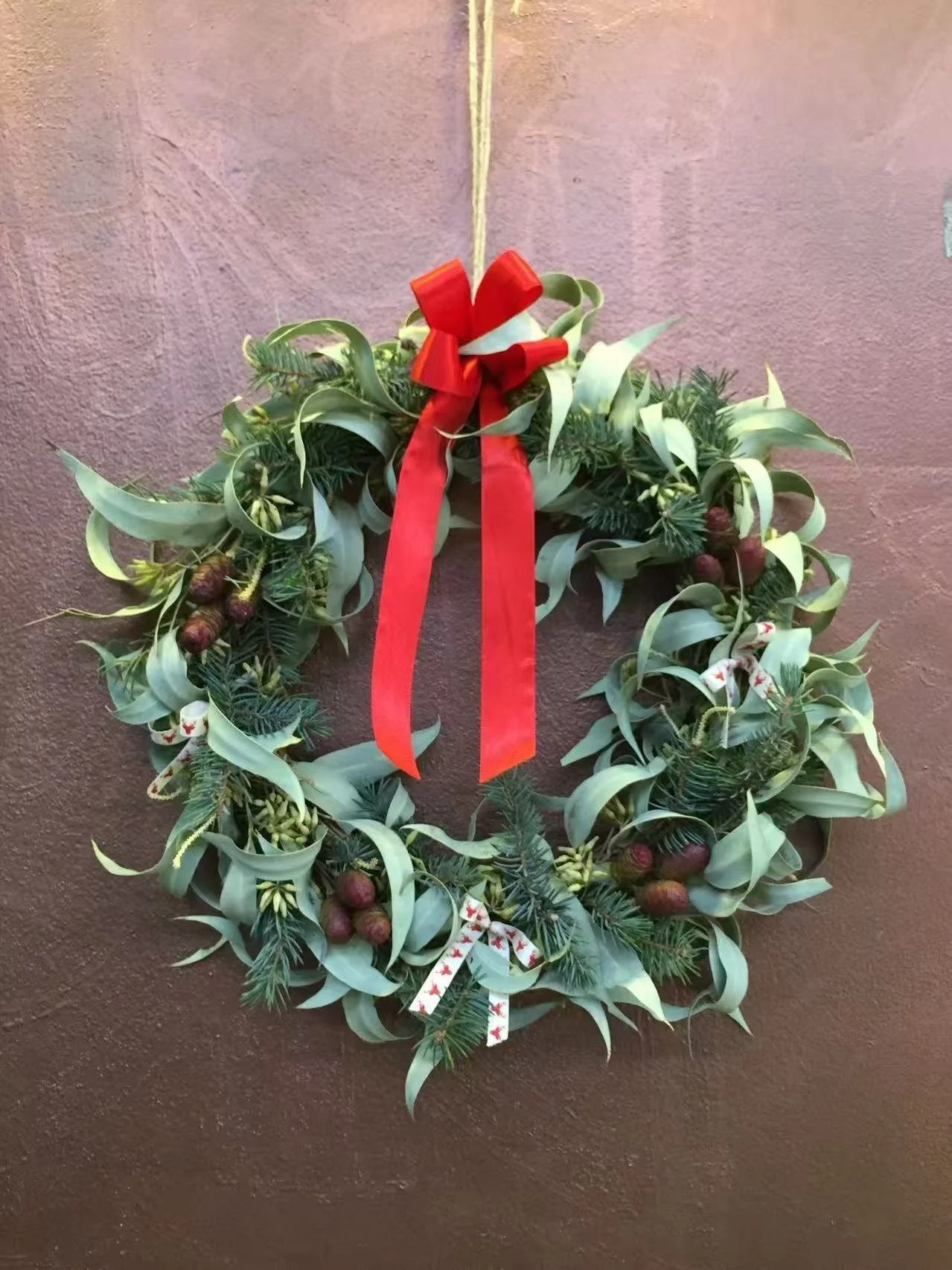 Christmas wreath decorated with green foliage, small pinecones, and white flowers, adorned with a red bow and hanging ribbon, hung on a brown wall.