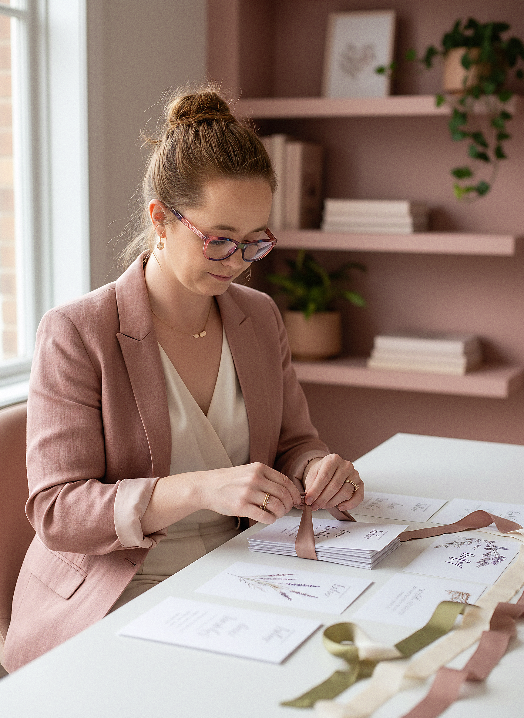 A woman in glasses and a pink blazer is sitting at a table, wrapping and tying ribbons around cards, possibly for an event or invitation.