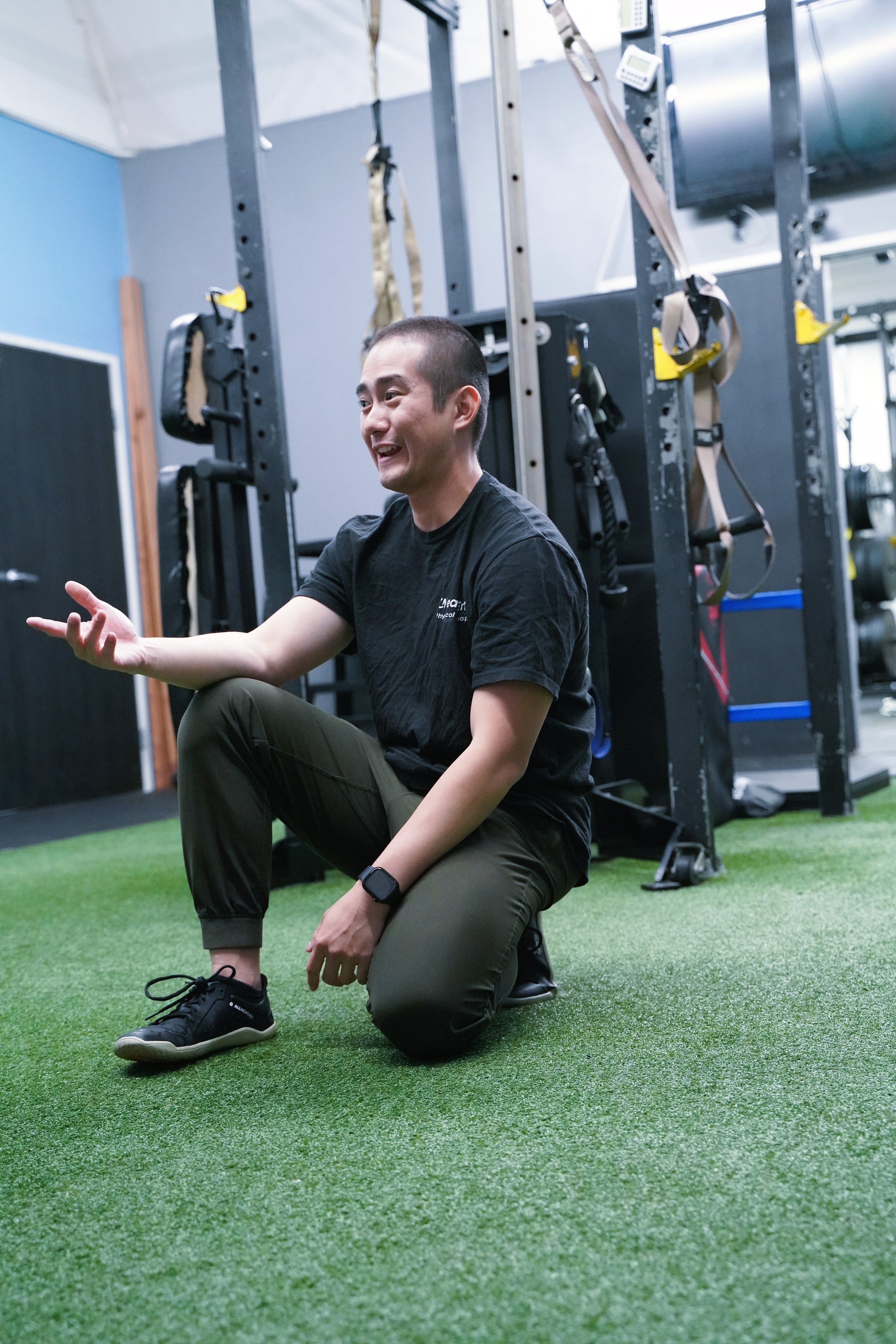 A physical therapist Dr. Chris Nguyen, sitting on the green gym floor in a fitness studio, gesturing with his left hand, smiling, with workout equipment in the background.
