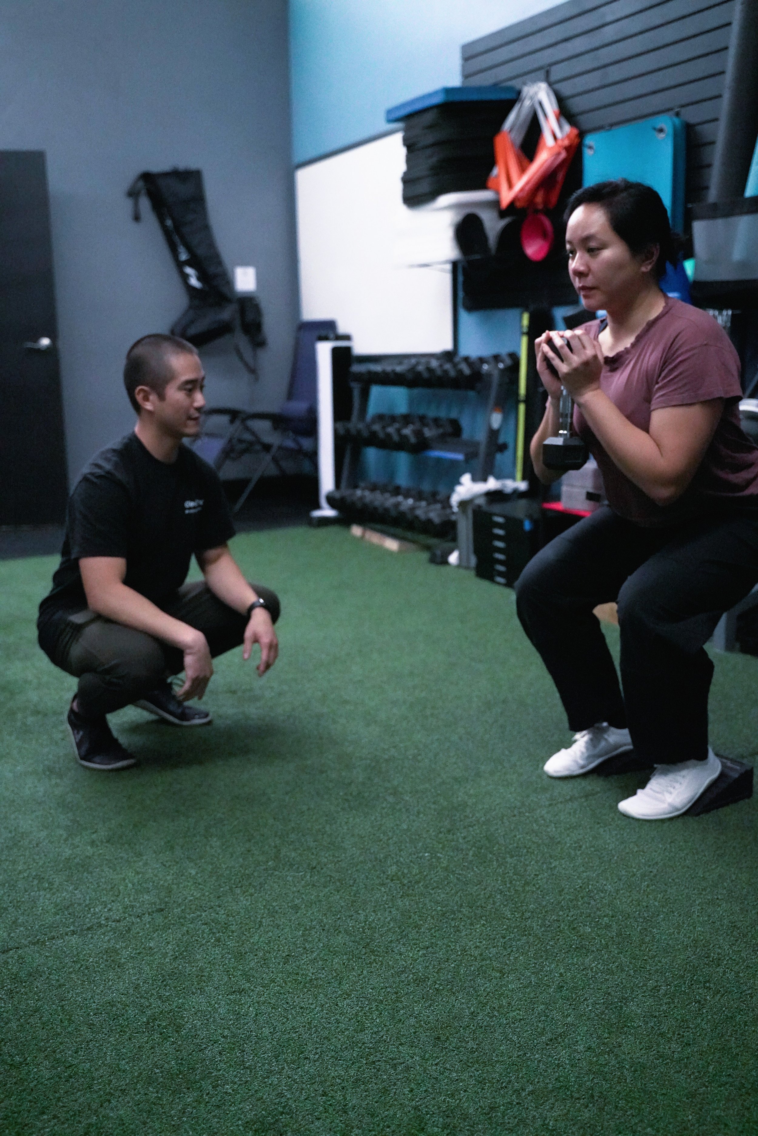 A woman exercises with a dumbbell while a trainer crouches down to instruct her in a gym with green turf flooring and various fitness equipment. She is dealing with an ACL sprain and is recovering for her physical therapist by doing squats.