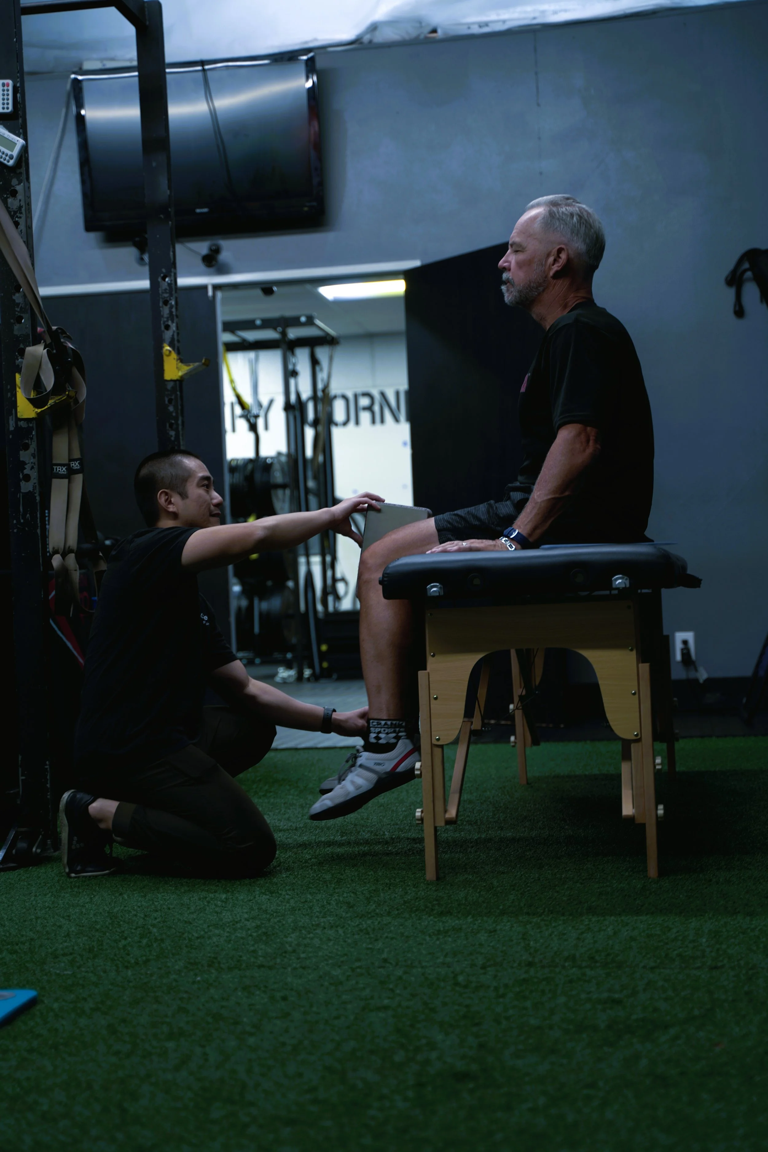 A man with gray hair and beard sitting on a therapy table while a trainer kneels in front of him and holds his ankle during a physical therapy session in a gym. They are helping to improve his hip mobility and osteoarthritis.