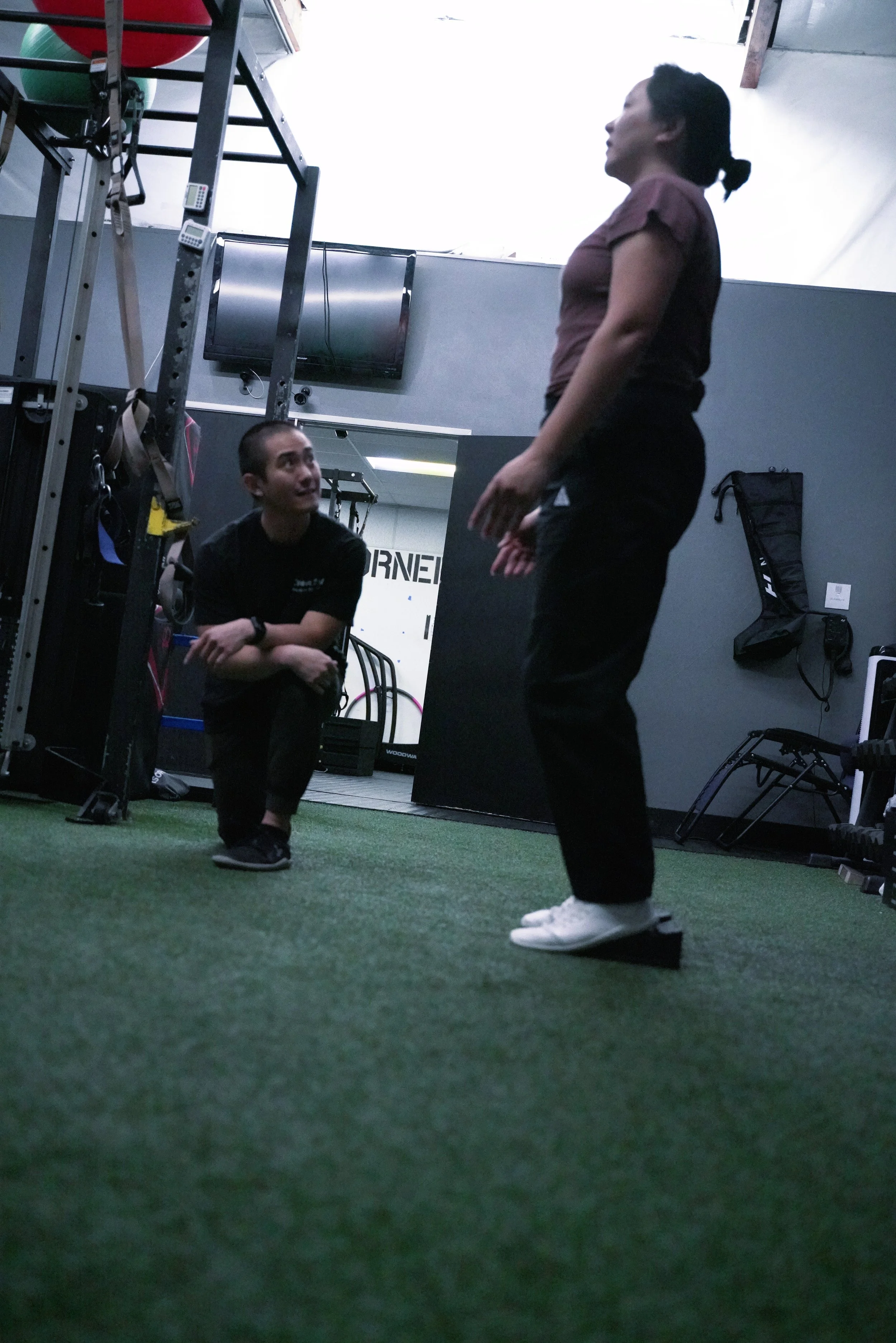 A trainer instructs a woman in a gym, with the woman standing and the trainer crouching on the dog somber in front of her, both engaging in a fitness or personal training session.