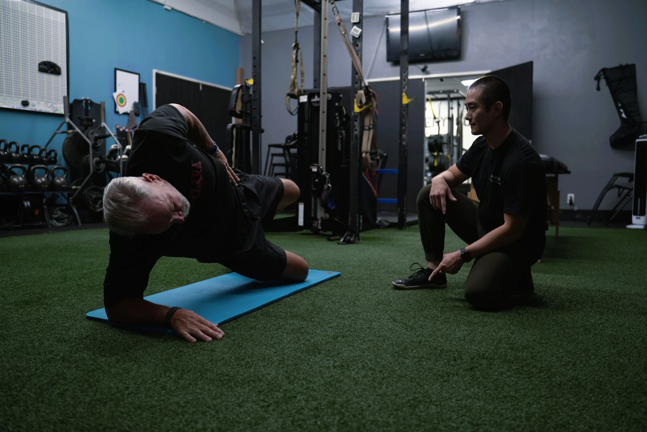 A man with gray hair and a beard is performing a side plank exercise on a blue mat in a gym, while a trainer kneels beside him, holding his ankle for support.