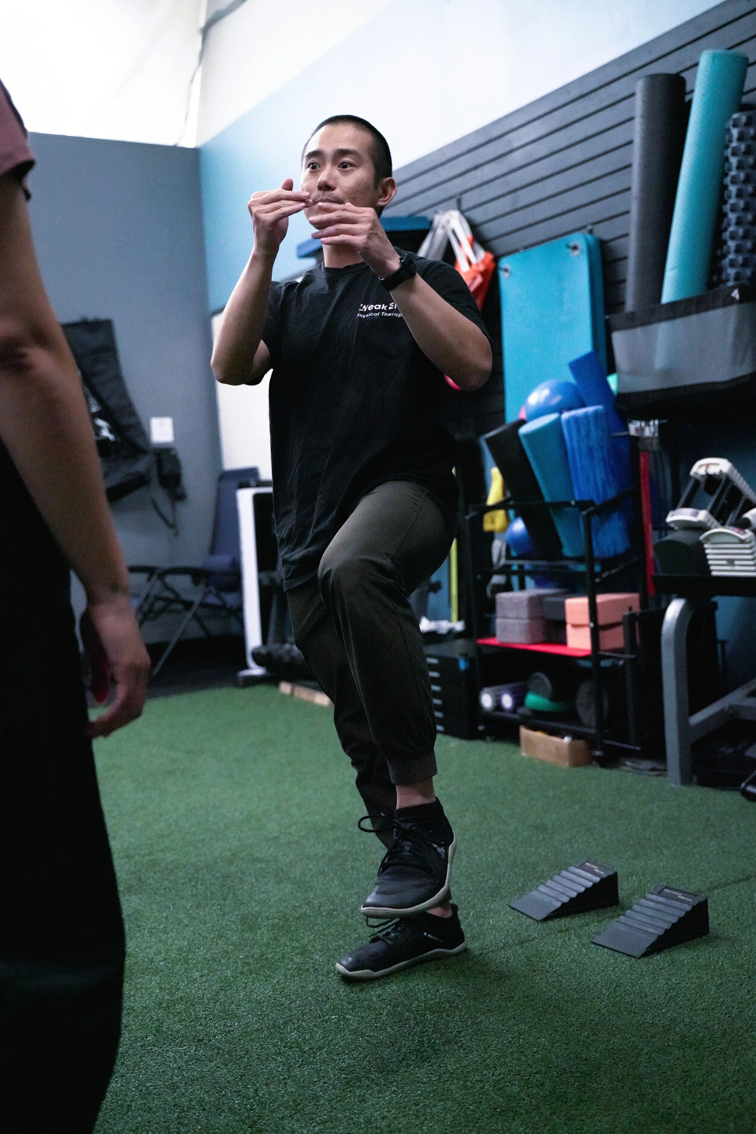 A man performing a high knee exercise in a gym, wearing black athletic clothing and sneakers, surrounded by workout equipment and mats.