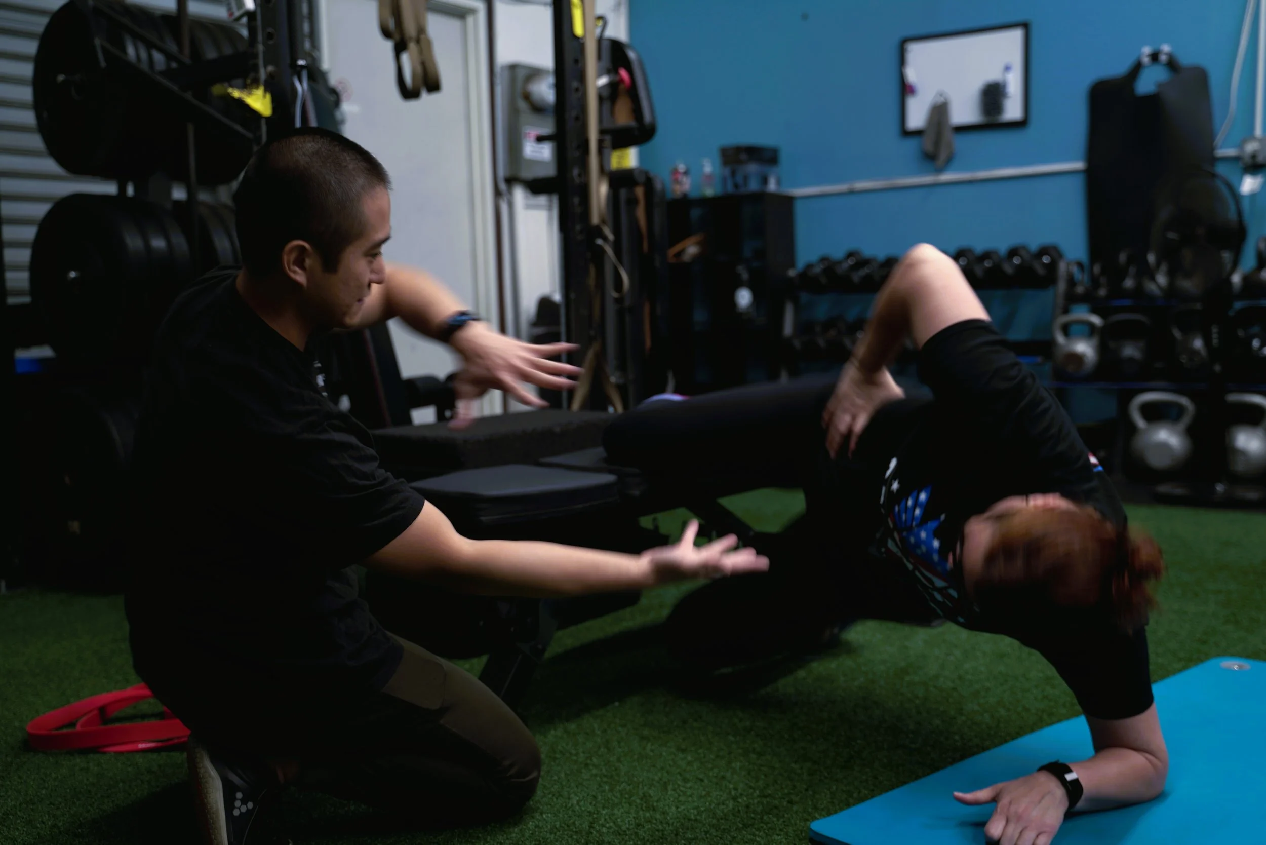 Personal trainer assisting a woman in a plank exercise at a gym, with kettlebells and gym equipment in the background.