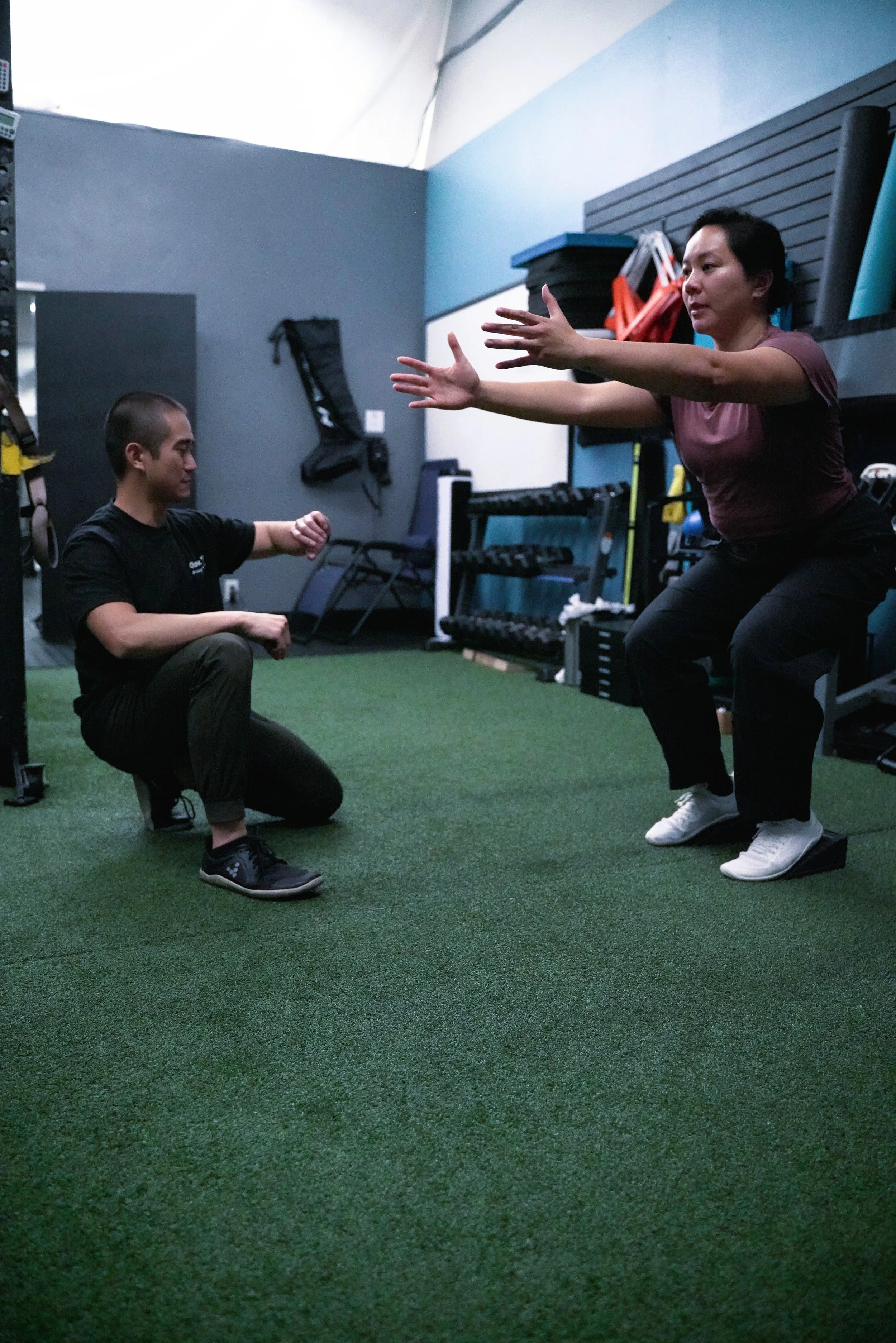 Personal trainer helping a client perform squats in a gym, with equipment and fitness gear in the background.