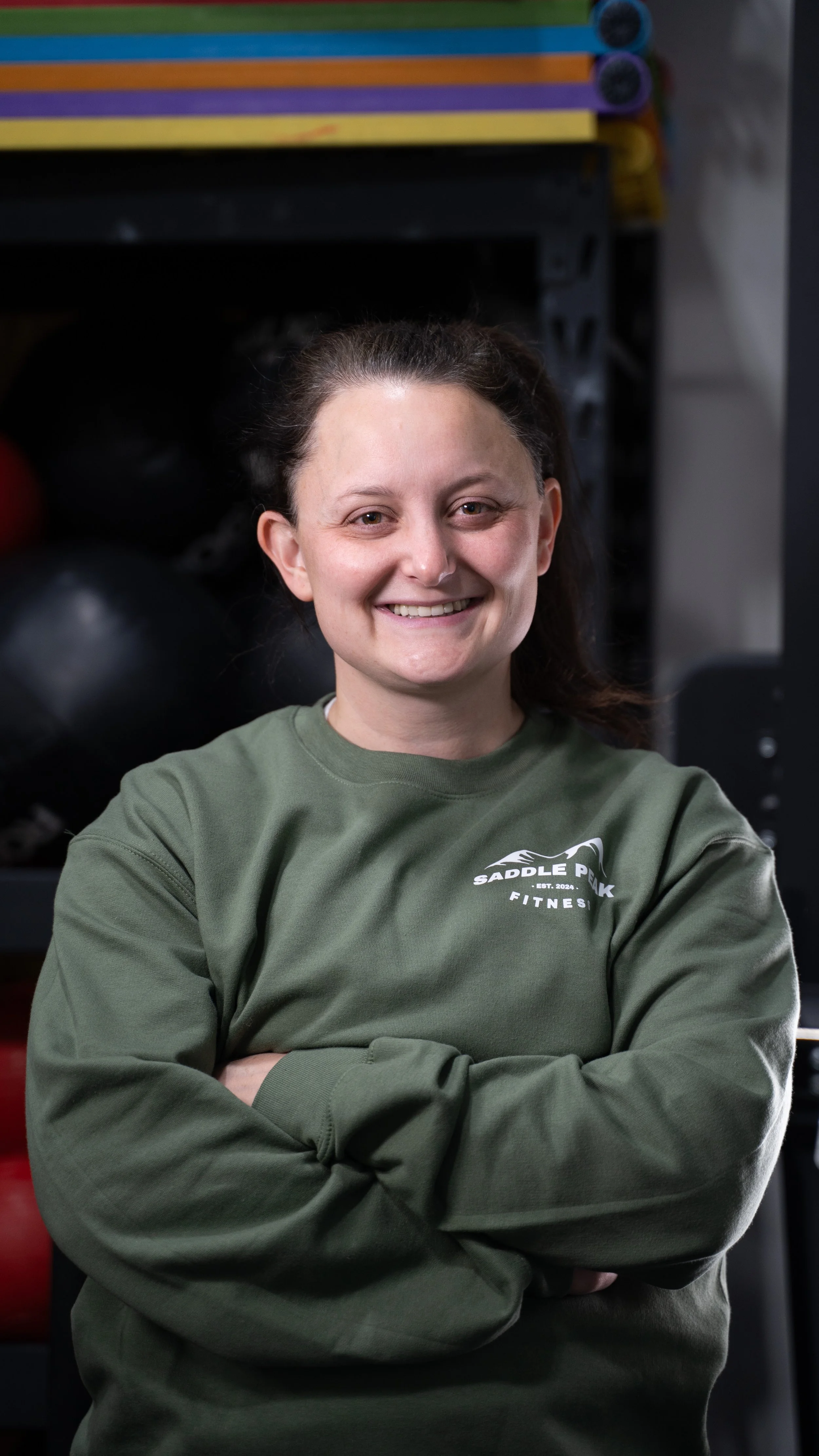 A woman in a green sweatshirt with crossed arms, smiling, in a gym setting with black kettlebells and colorful mats in the background.