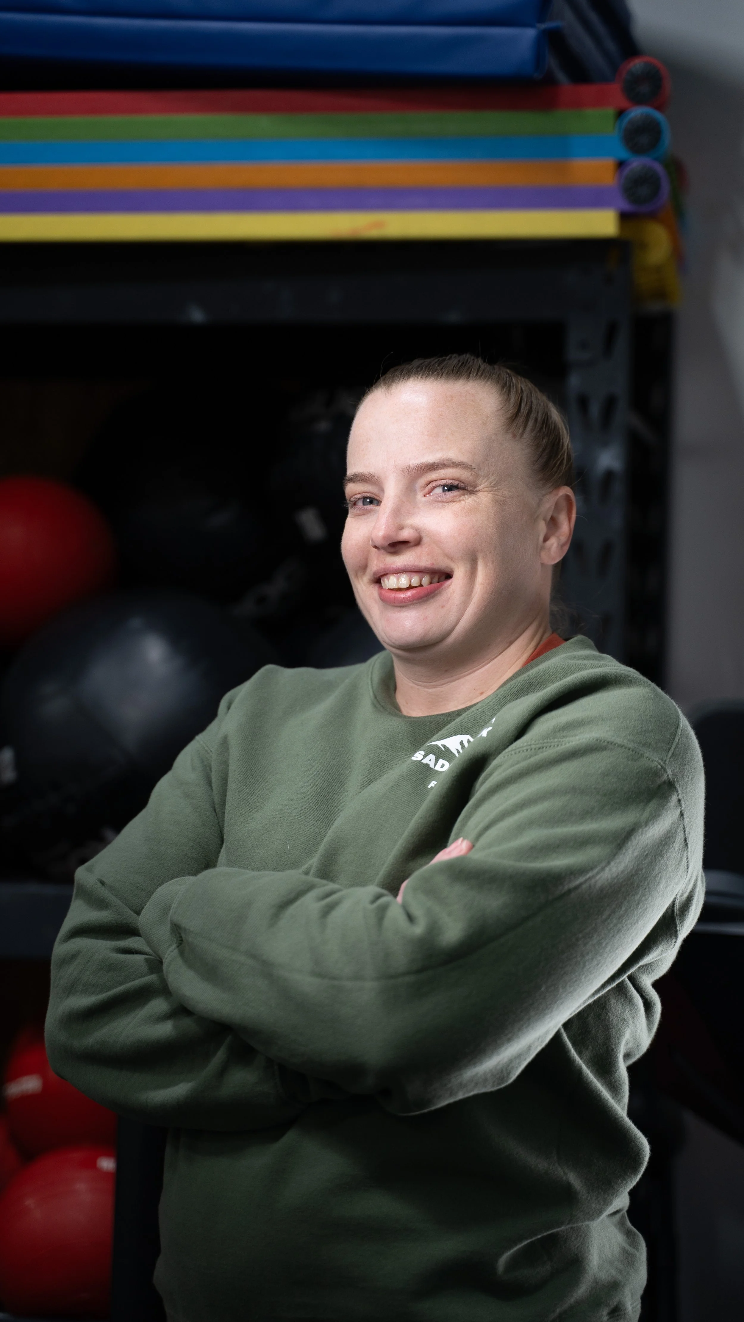 A smiling woman with light skin and red hair pulled back, wearing a green sweatshirt with a logo, standing with her arms crossed in a gym environment with exercise equipment in the background.