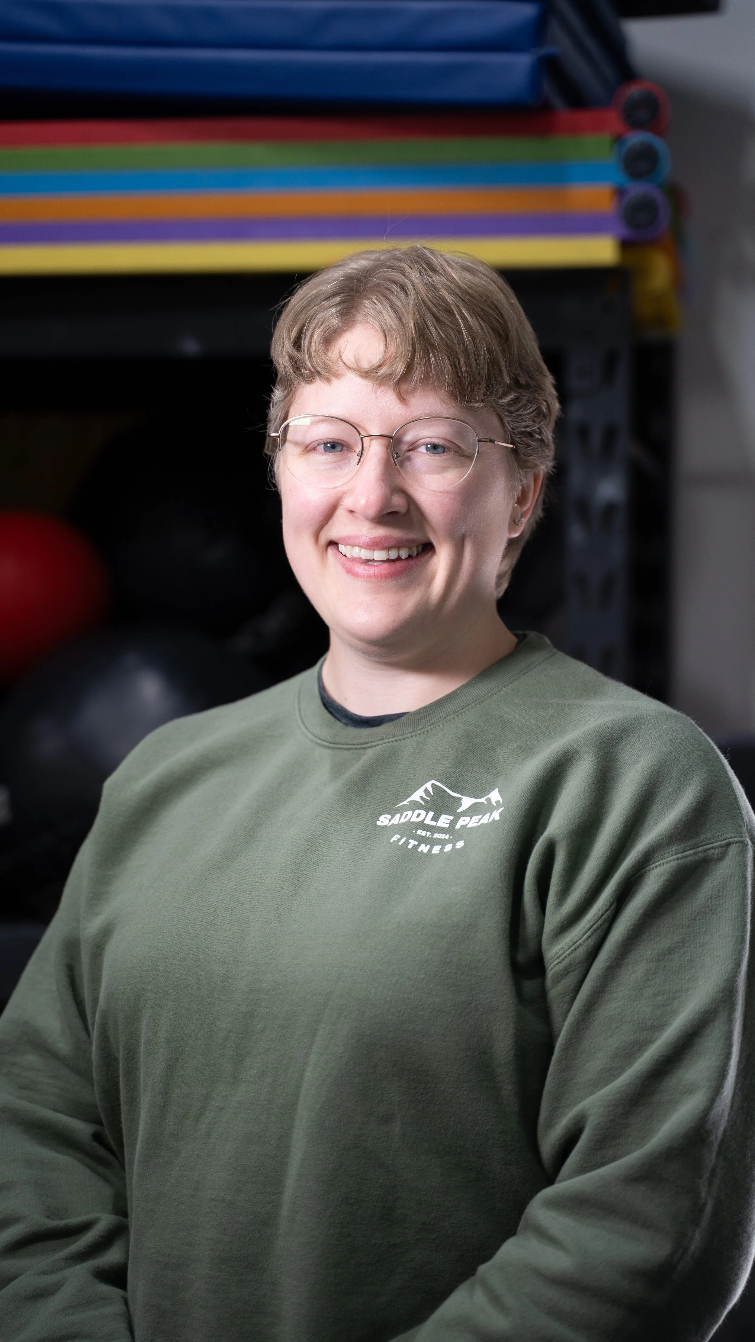 A smiling person with short light brown hair and glasses, wearing a green sweatshirt with a logo that says 'Saddle Peak Fitness,' sitting in a gym with black and red gym balls and shelves of exercise mats in the background.