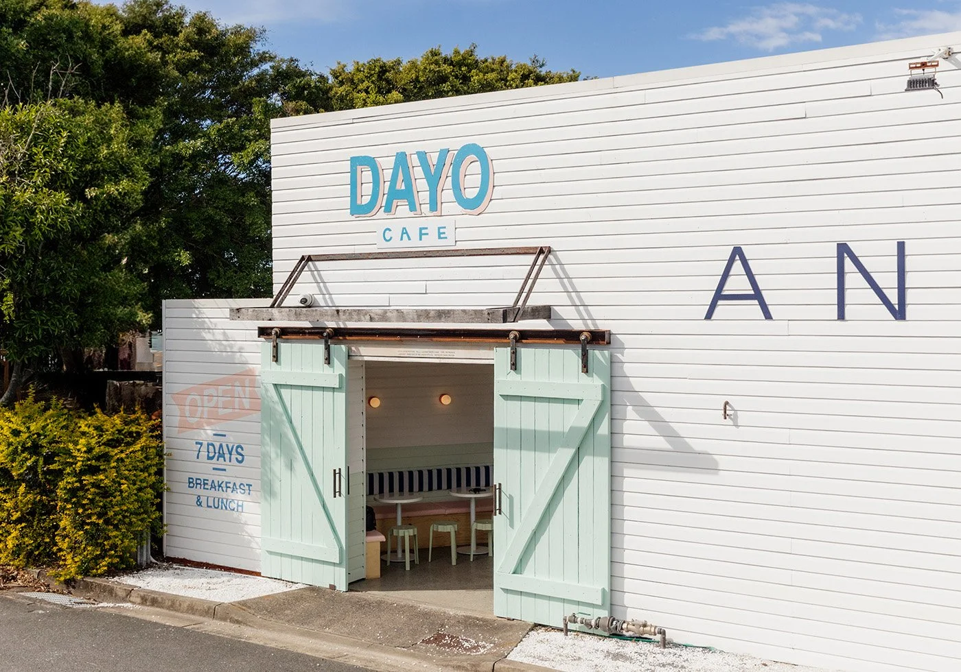 Exterior of a white cafe building with blue and navy lettering, including 'Dayo Cafe' and 'Open 7 Days Breakfast & Lunch.' Light green barn-style doors are open, revealing a seating area inside.