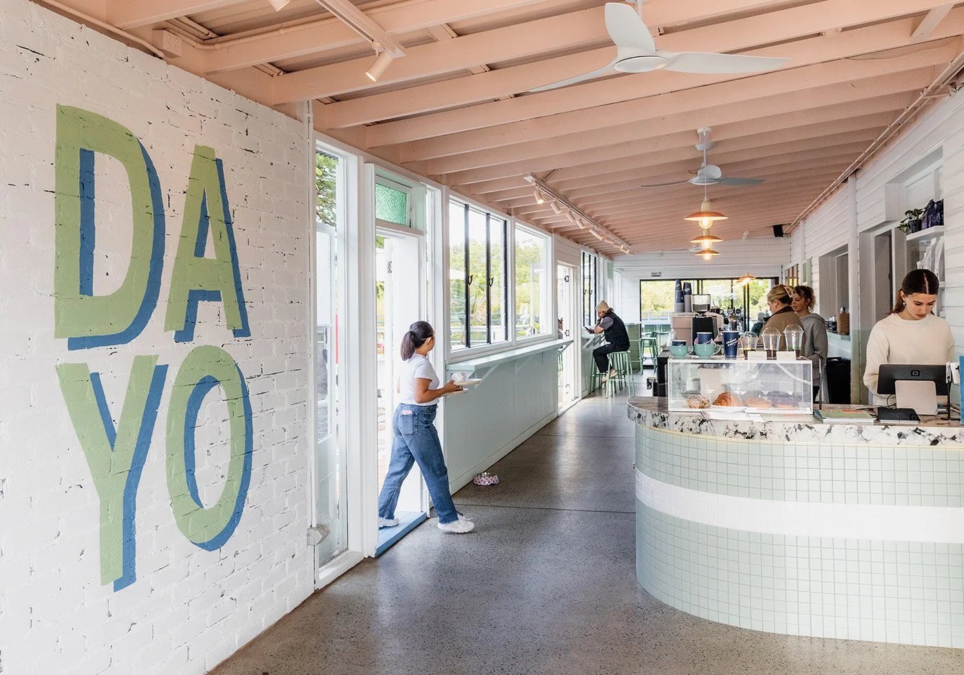 Interior of a bright, modern cafe with white brick walls, large windows, and a curved marble counter. Customers and staff are inside, and a large pastel painted sign on the wall reads 'DAYO'.