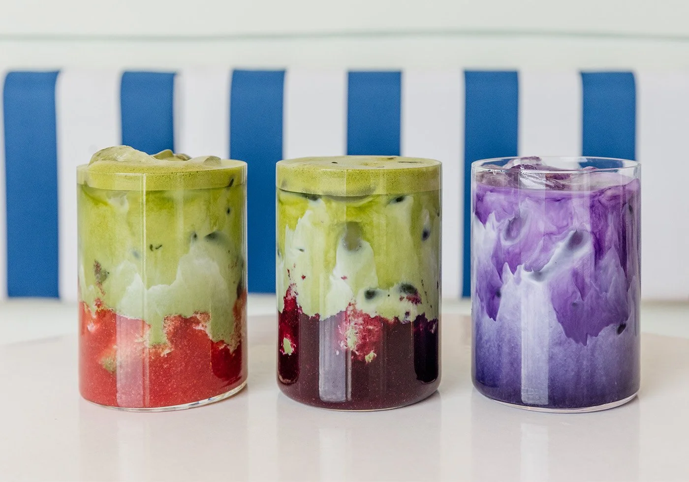 Three colorful layered drinks with whipped foam on top, placed on a white surface with a striped blue and white background.