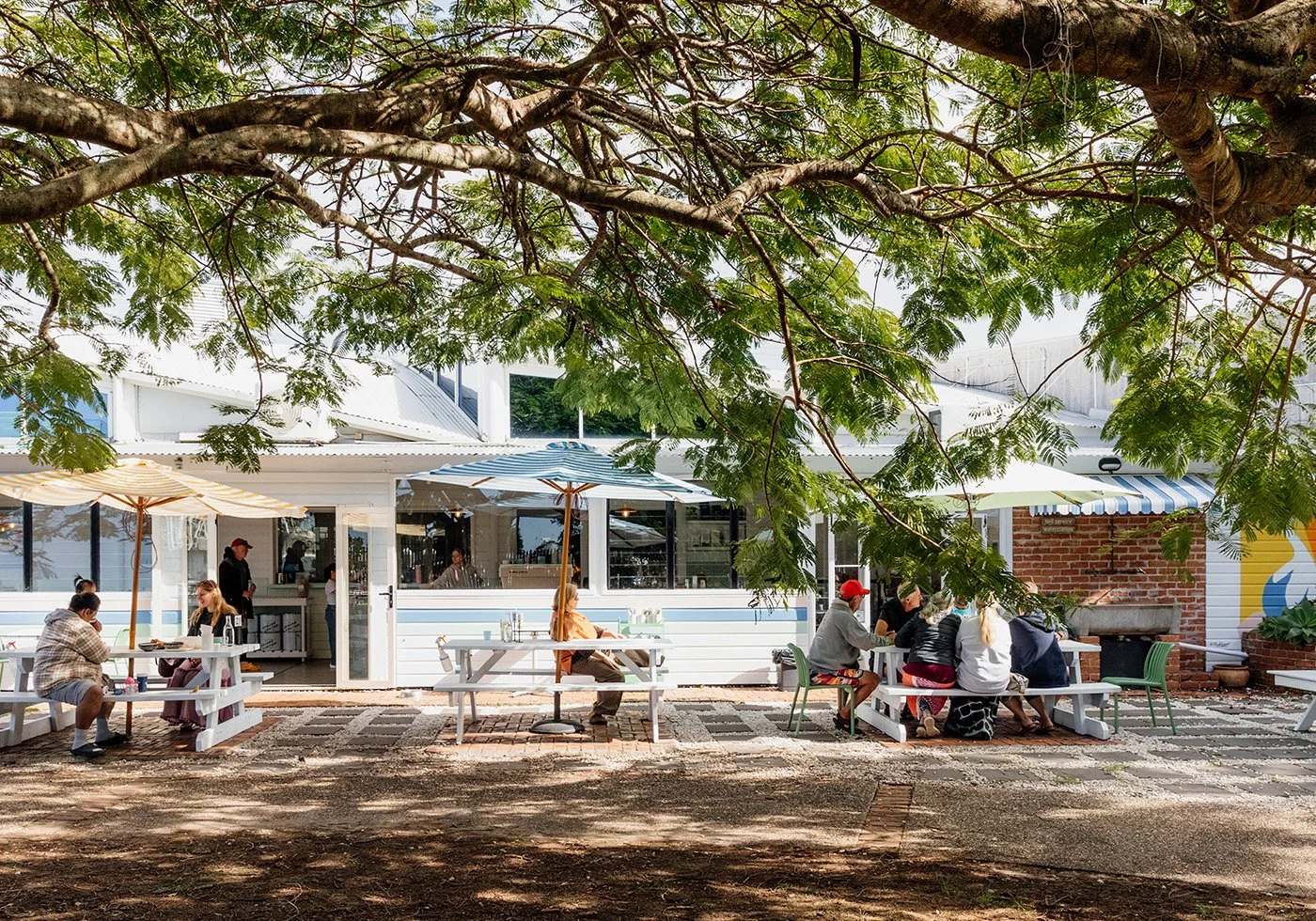 Outdoor patio at a cafe with several groups of people sitting at tables under large umbrellas, surrounded by trees and greenery.