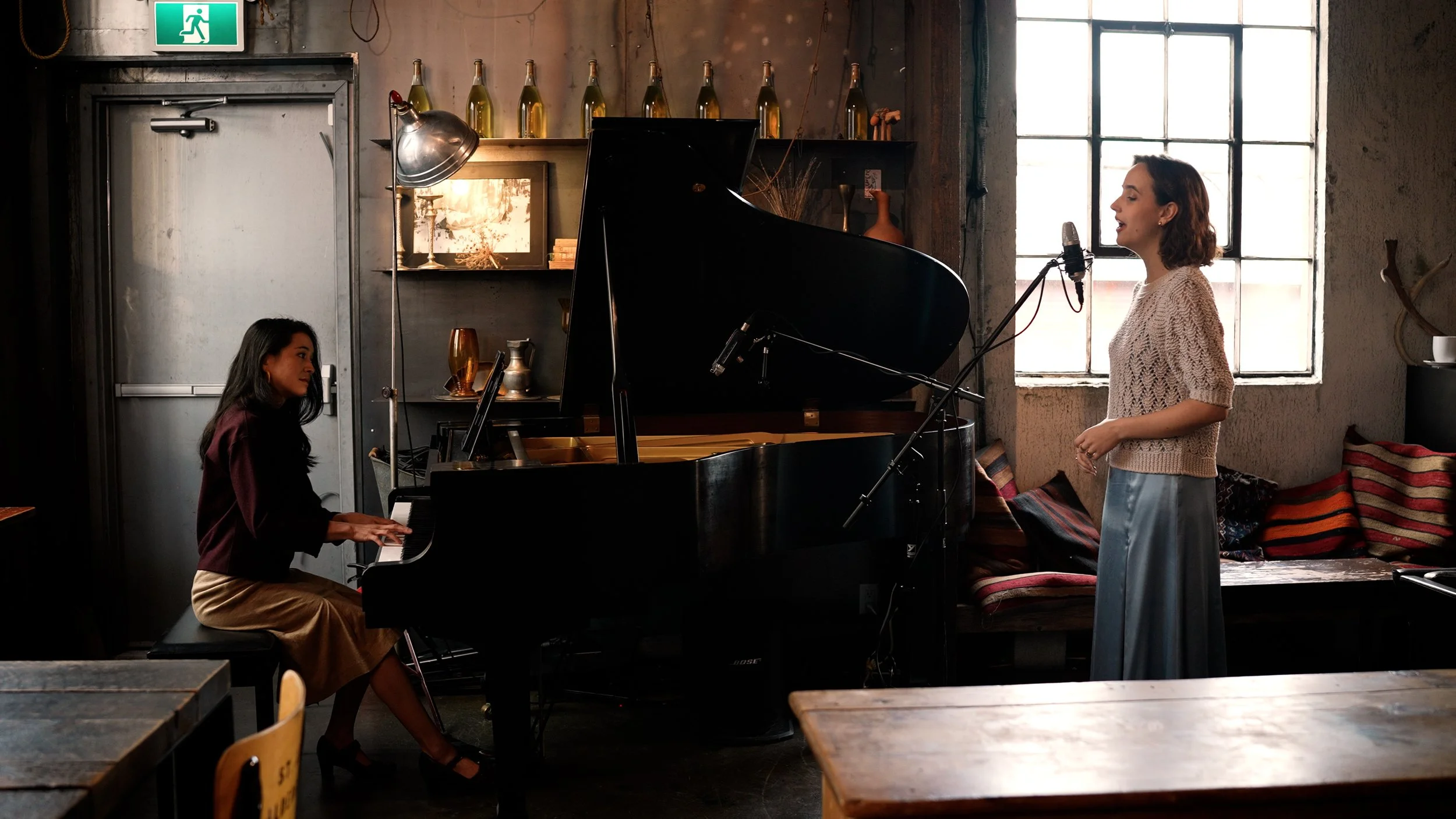 A woman singing into a microphone while a woman plays piano in a cozy, rustic room with large window and warm decor.