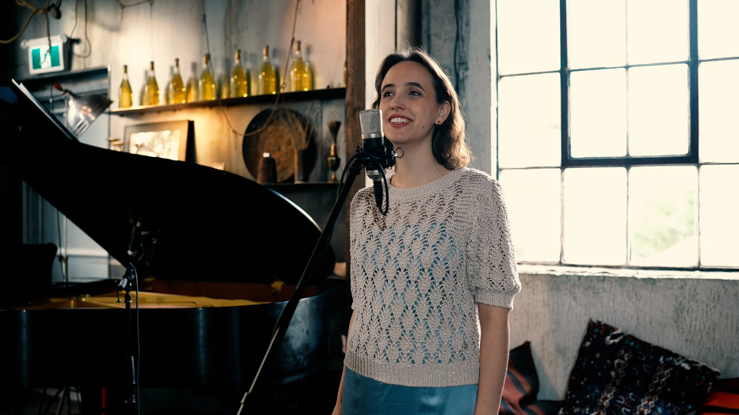 A woman singing into a microphone in Fabrique St George, with a grand piano and large window letting in natural light.