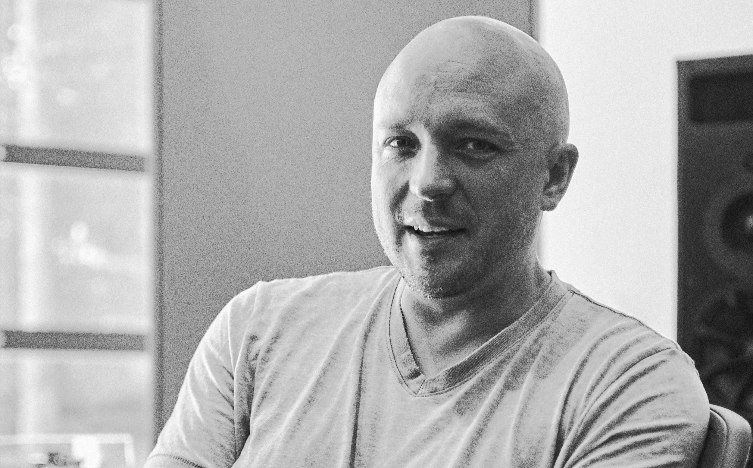 A black and white photo of a man wearing a light T-shirt, sitting in a room with a speaker in the background.